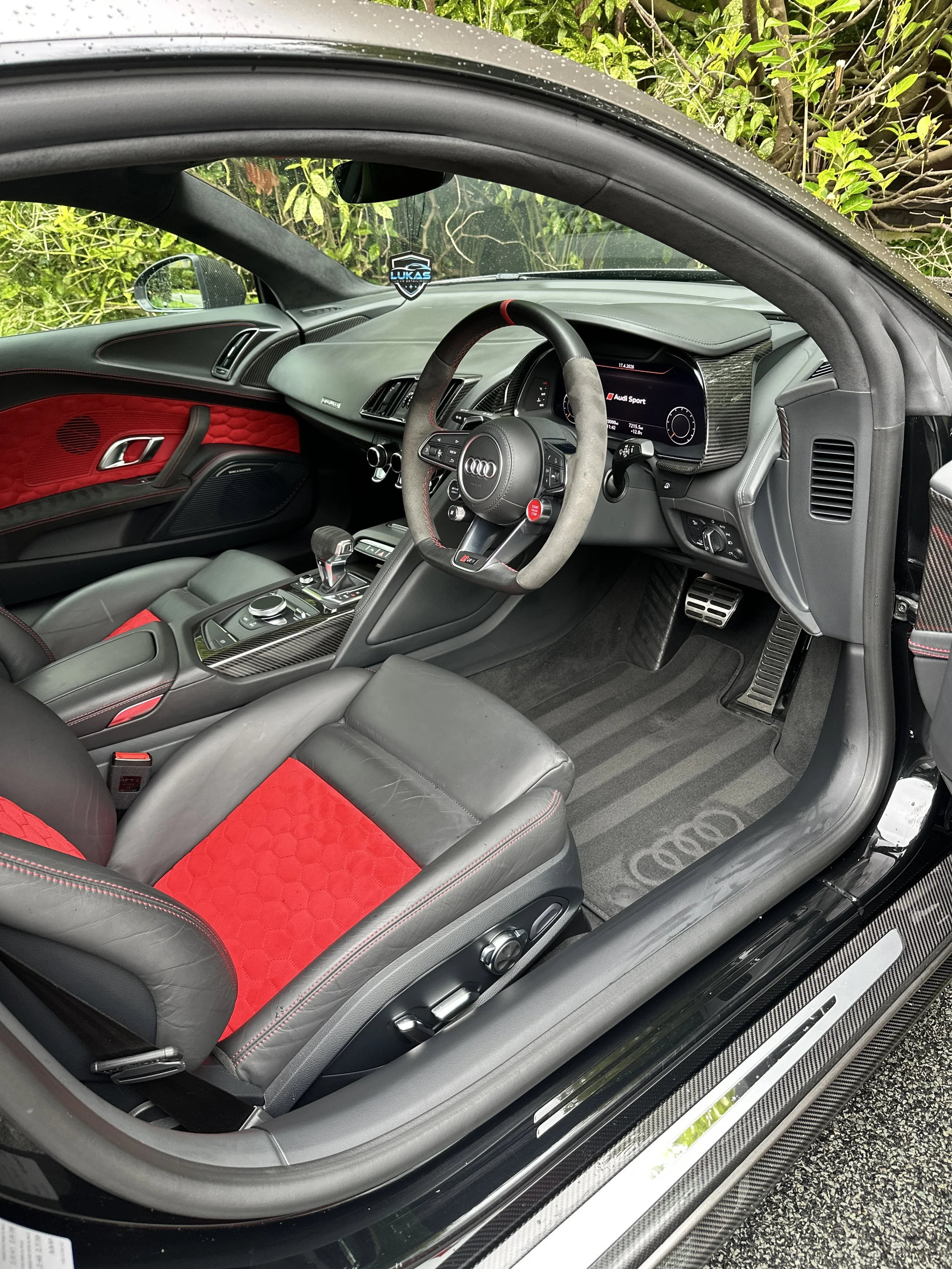 The interior of a black Audi sports car, featuring a black dashboard with a digital display, a racing steering wheel with the Audi logo and red accents, black leather seats with red quilted inserts, and carbon fiber trim on the center console and door panels. The car is parked outside amid green foliage.
