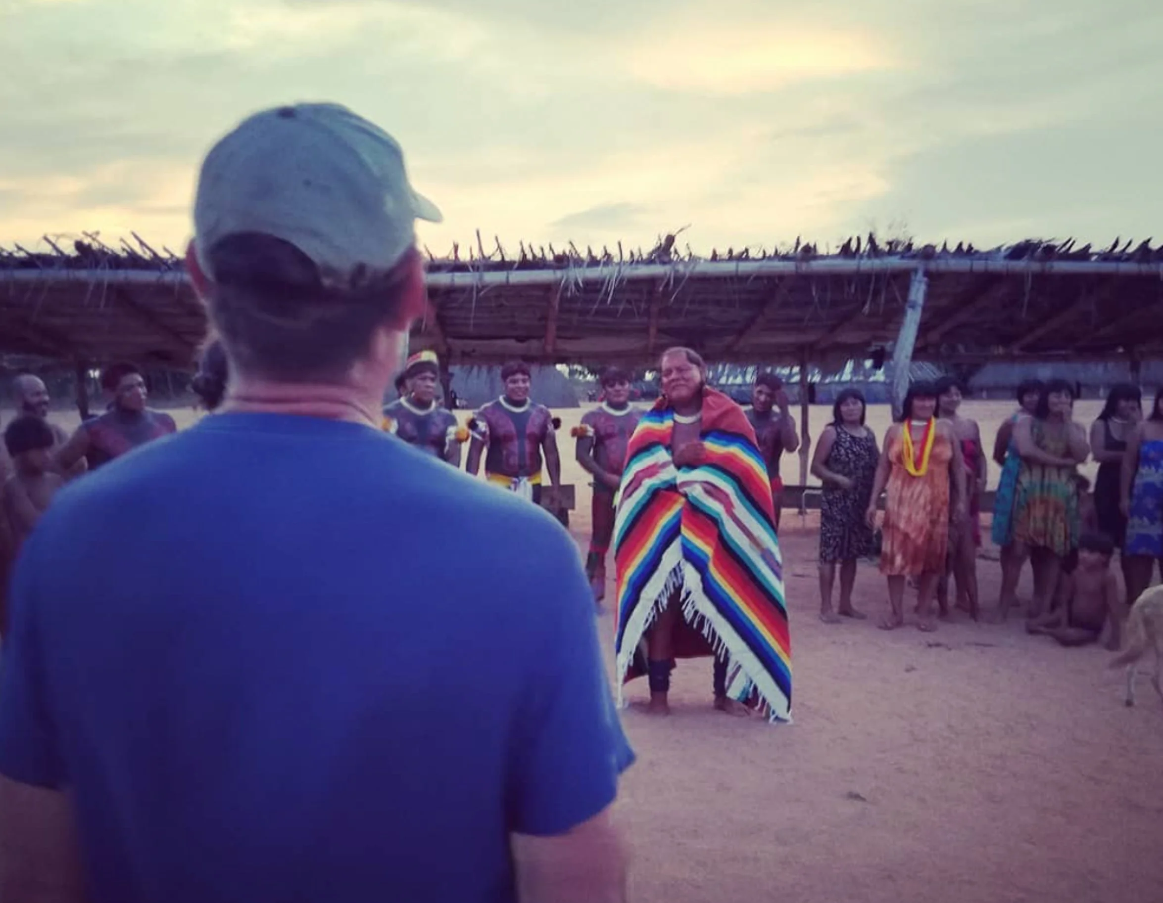 Homem de costas, usando boné, observando um grupo de indígenas com trajes coloridos, sob uma estrutura de capim, ao entardecer.