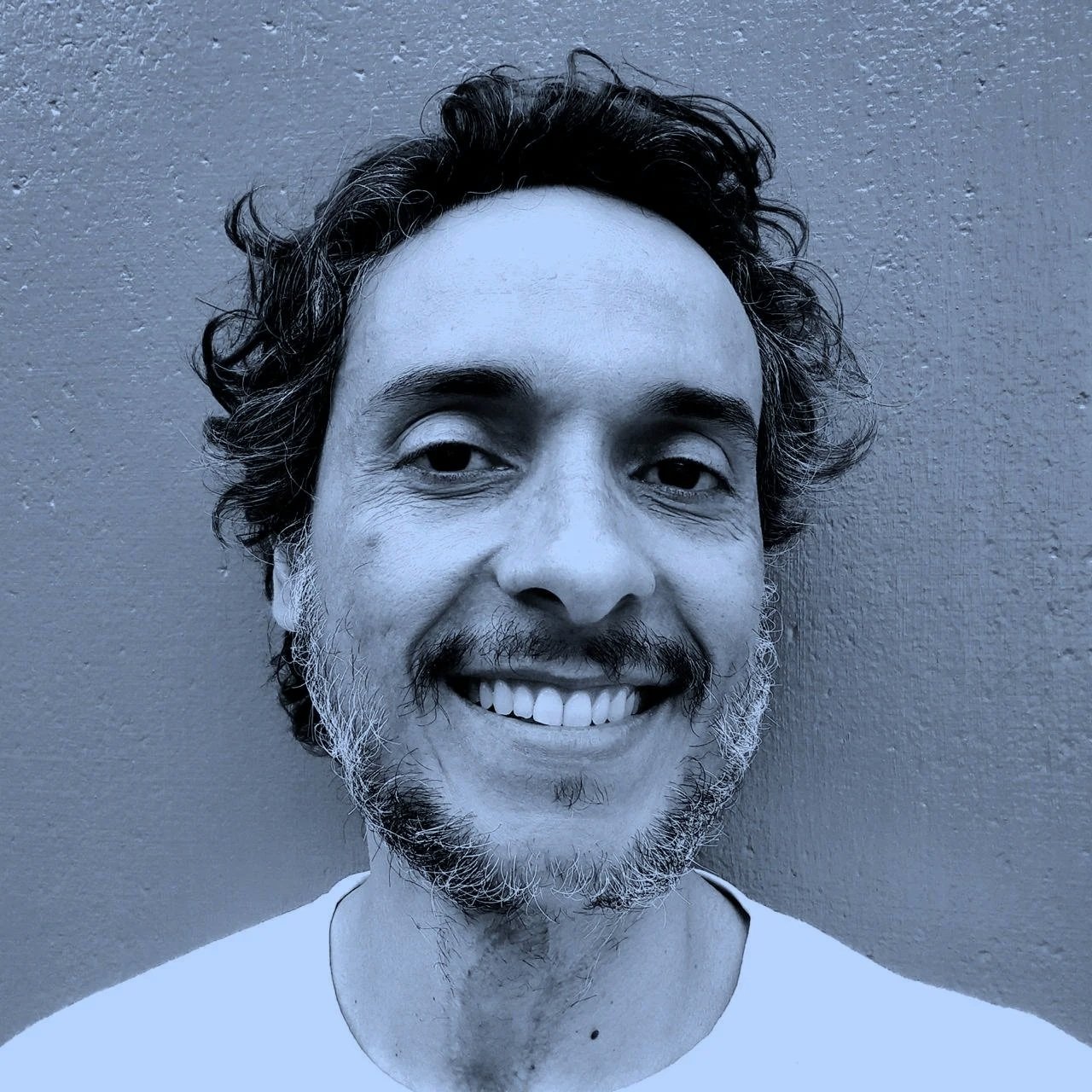 Retrato em preto e branco de um homem sorrindo, com cabelo e barba curtos, contra parede de textura lisa.
