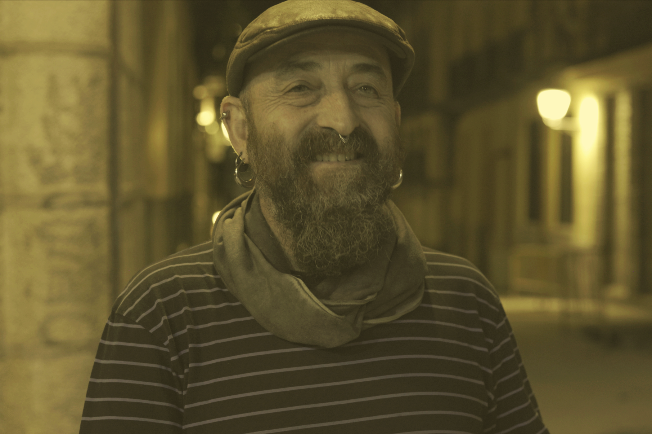 Homem sorridente com barba, usando chapéu e brincos, vestindo uma blusa listrada, em ambiente com iluminação dourada e fundo desfocado.