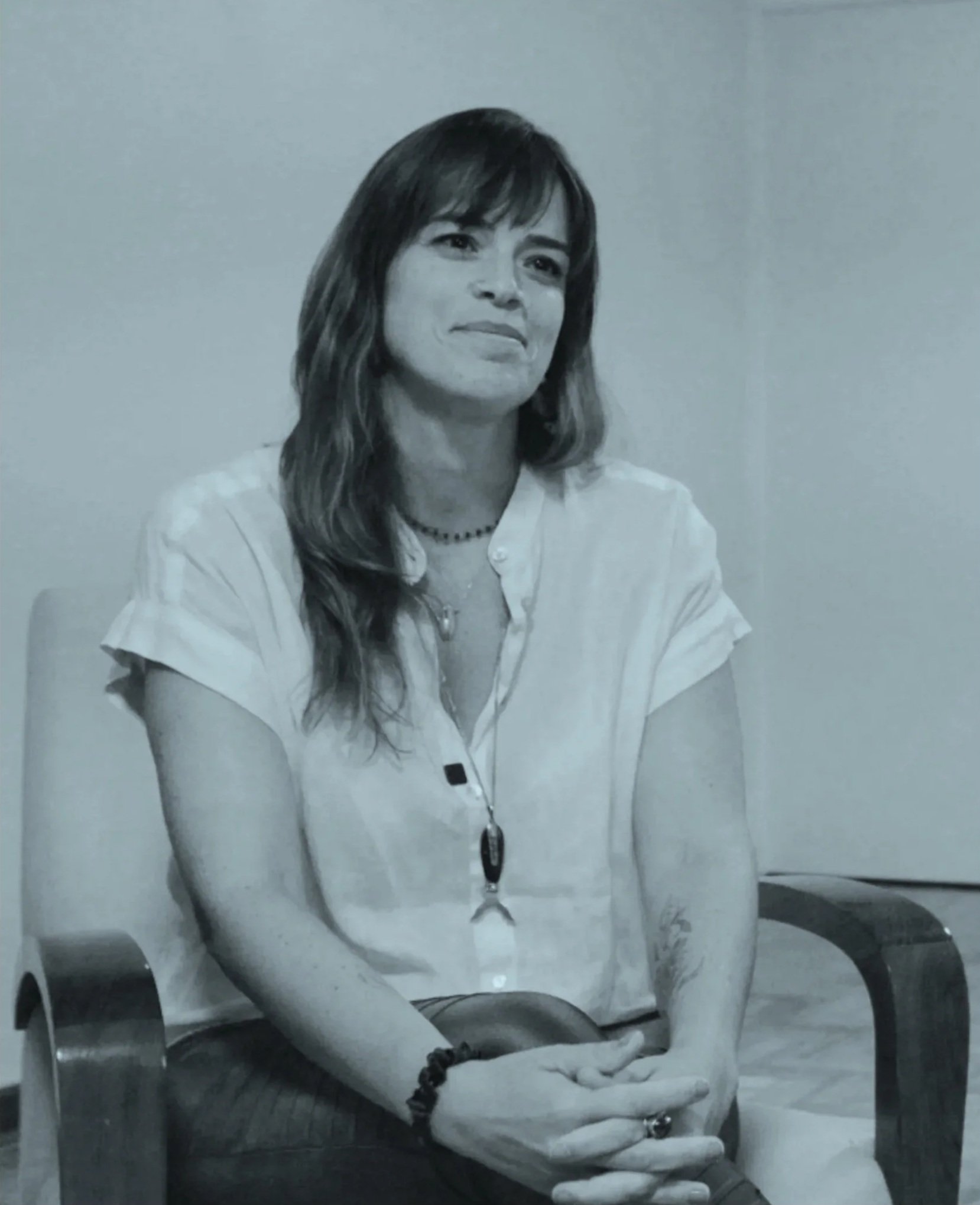 Mulher com cabelo longo e escuro sentado em uma cadeira de escritório, usando uma camisa branca, com um colar preto. Ela está olhando para frente e segurando um dispositivo móvel nas mãos. Ao fundo, uma parede clara e uma cadeira de escritório preta.