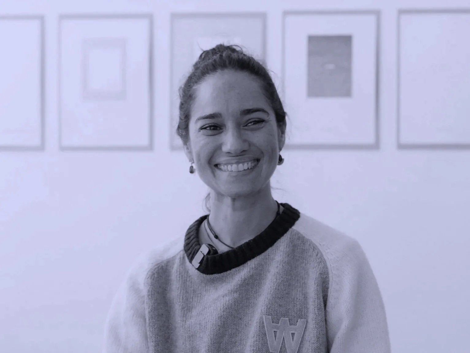 Retrato em preto e branco de uma mulher sorrindo, com cabelo preso, usando brincos, um suéter claro, um colar e um tecido no pescoço, em uma sala com quadros na parede ao fundo.