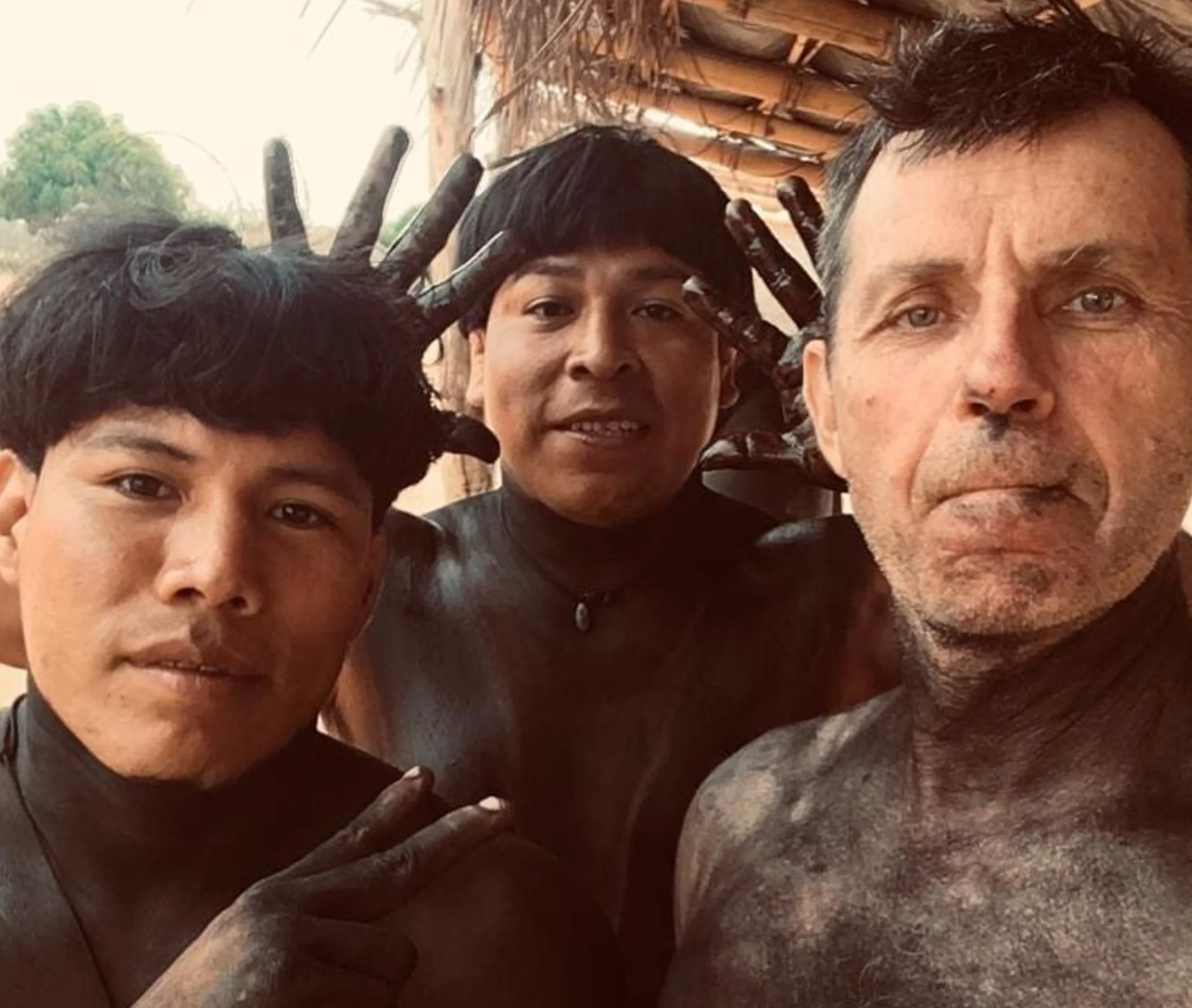 Grupo de quatro pessoas fazendo pose para a foto, incluindo duas pessoas de pele clara, uma de pele morena e uma de pele mais escura, com fundo de uma estrutura de madeira.