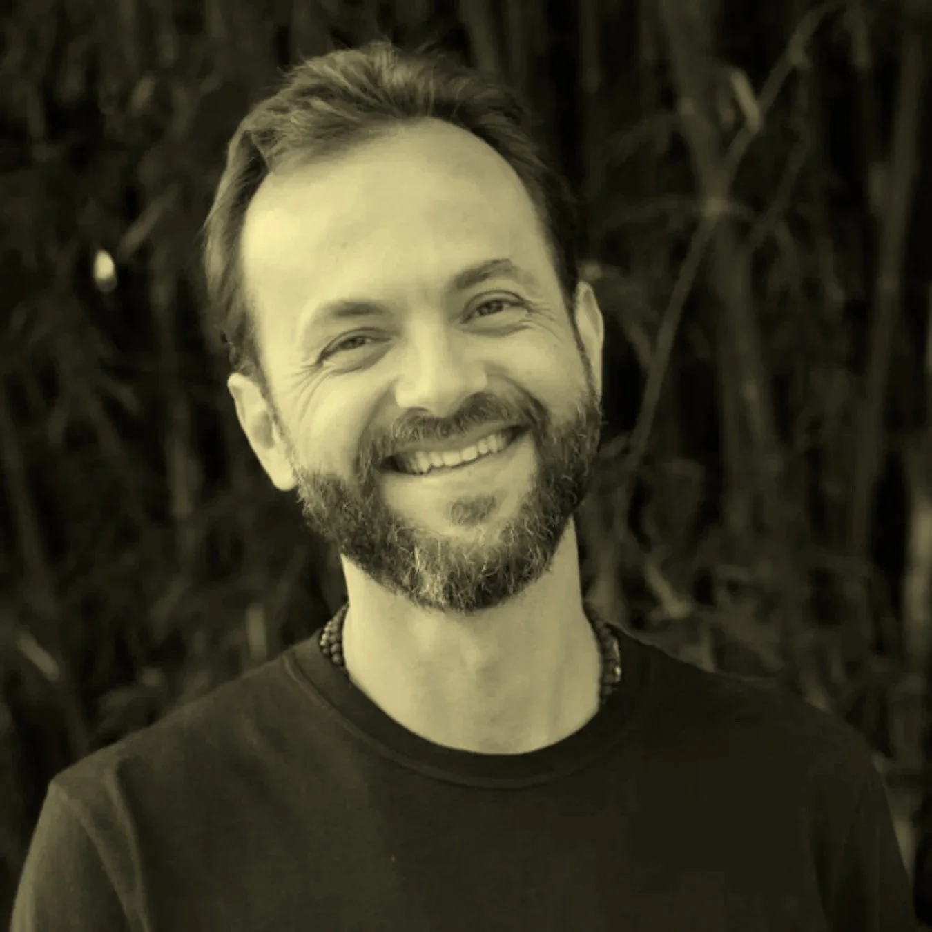 Retrato em sepia de um homem sorridente com barba, cabelo curto e camisa preta, contra fundo de vegetação.