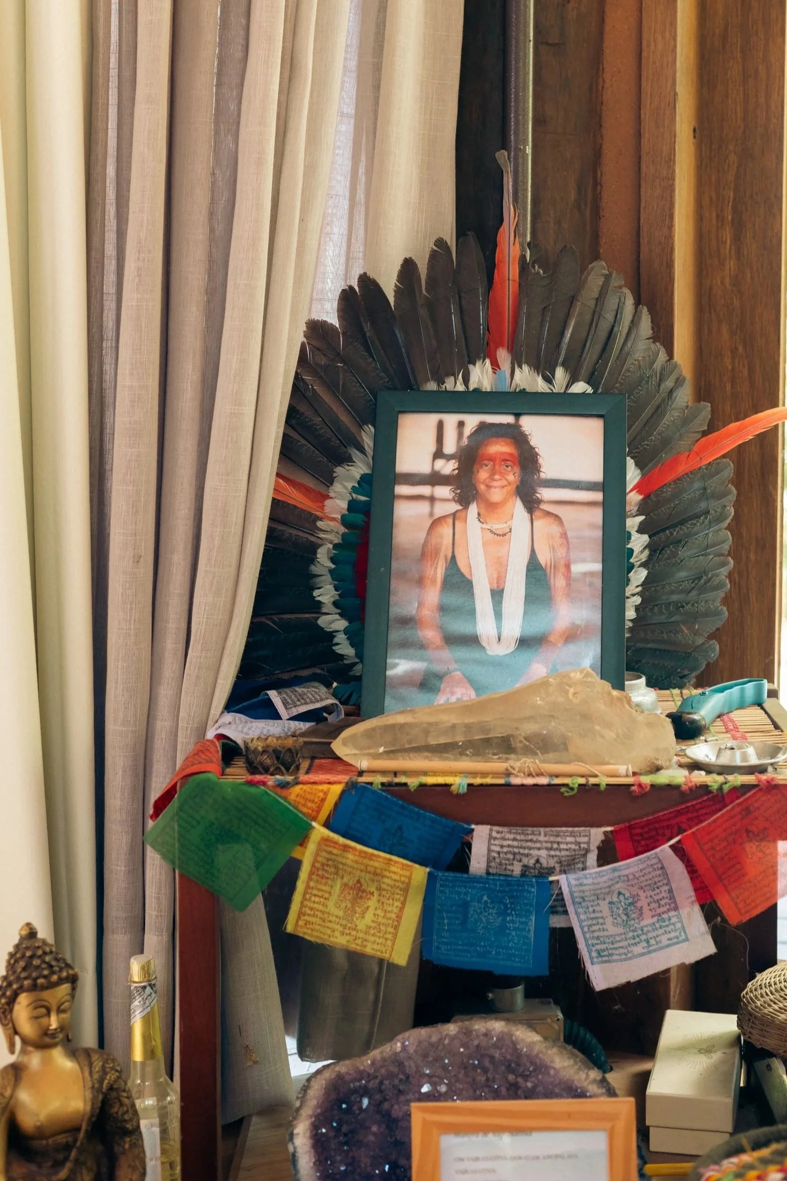Imagem de um altar com uma foto de uma pessoa sorridente, decorada com penas coloridas, mensagens e objetos diversos. Há uma vela, uma escultura de cabeça de Buda e pedras ao redor.