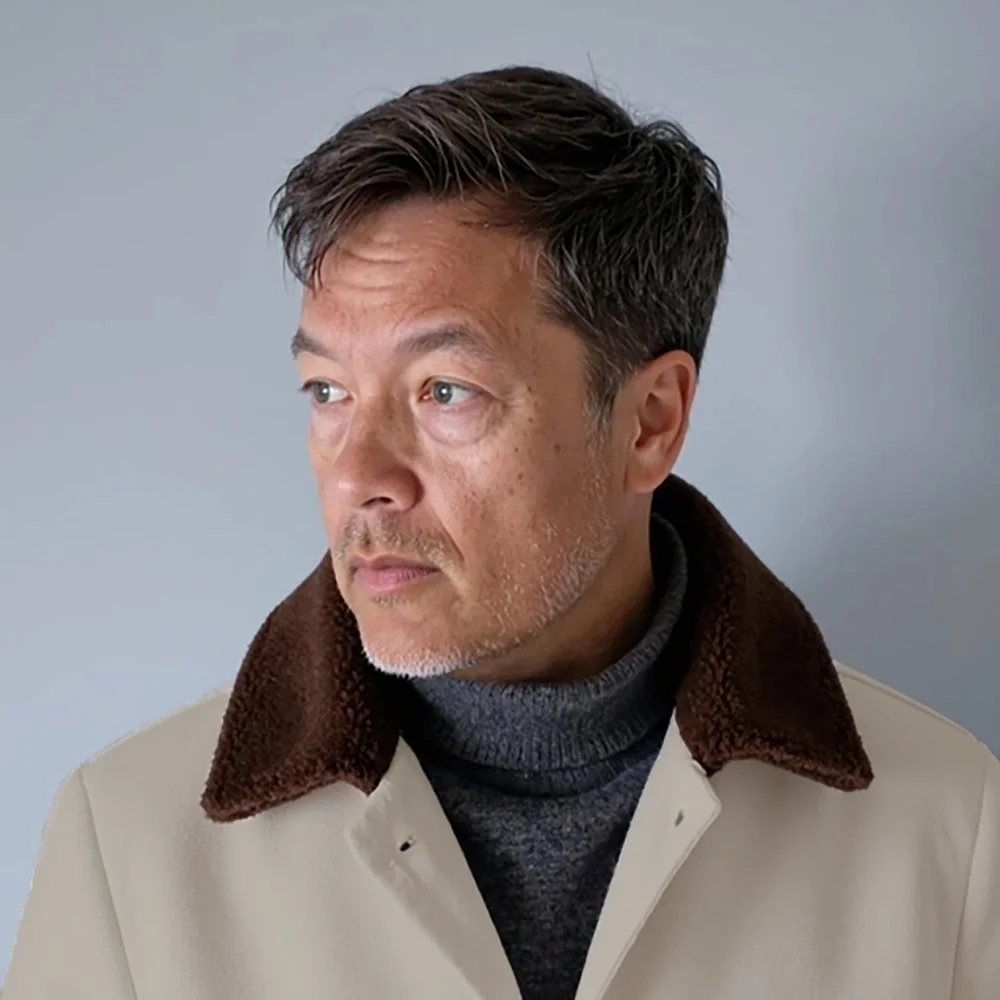 A middle-aged man with short dark hair and stubble, wearing a beige coat with a brown collar and a gray turtleneck, looking to his right against a plain light gray background.