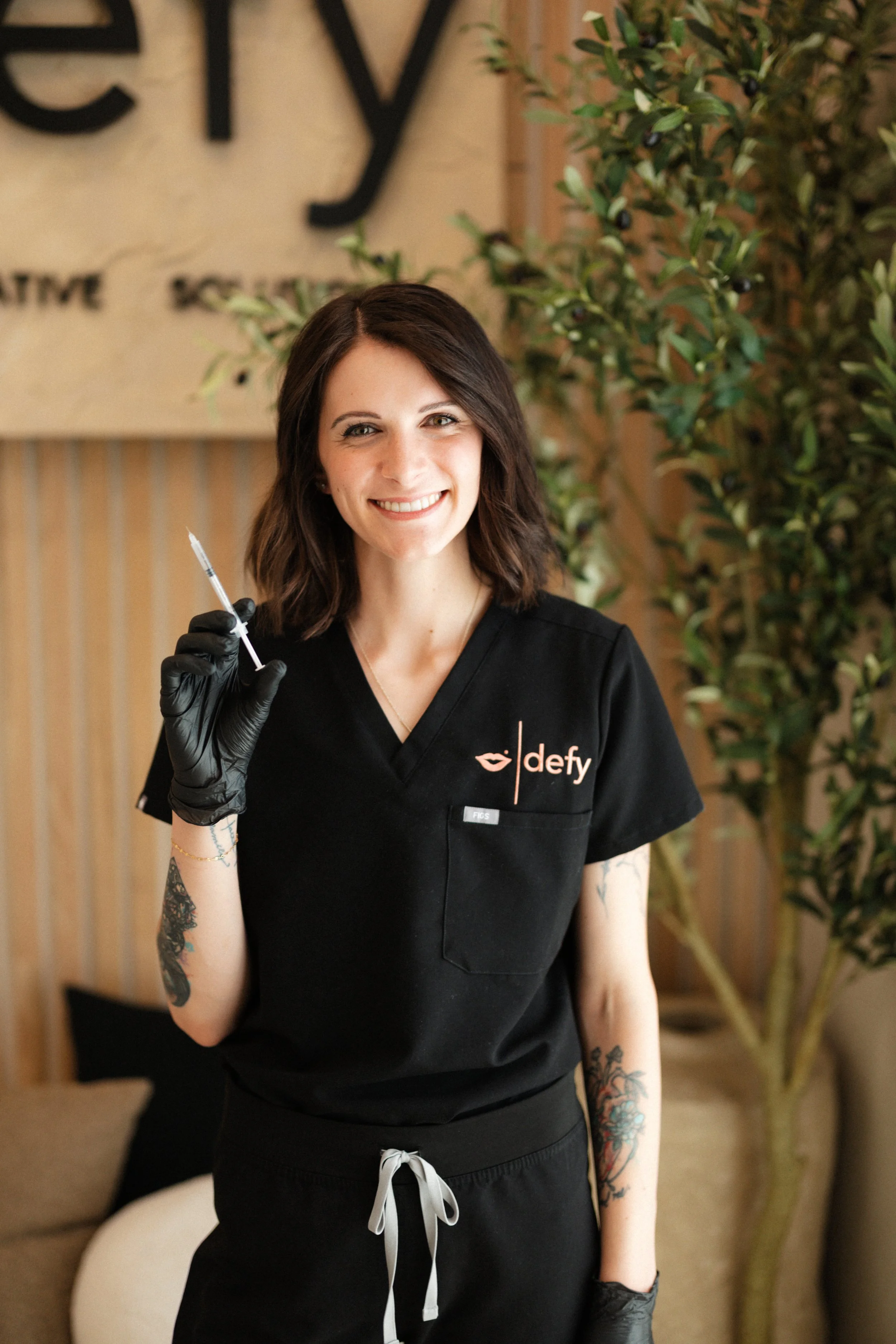 A smiling woman with dark brown hair and tattoos on her arms, wearing black scrubs and gloves, holding a syringe, standing indoors near a plant and wooden wall with a partially visible sign. Defy RS | Daxxify & Lip Filler in Pueblo West CO