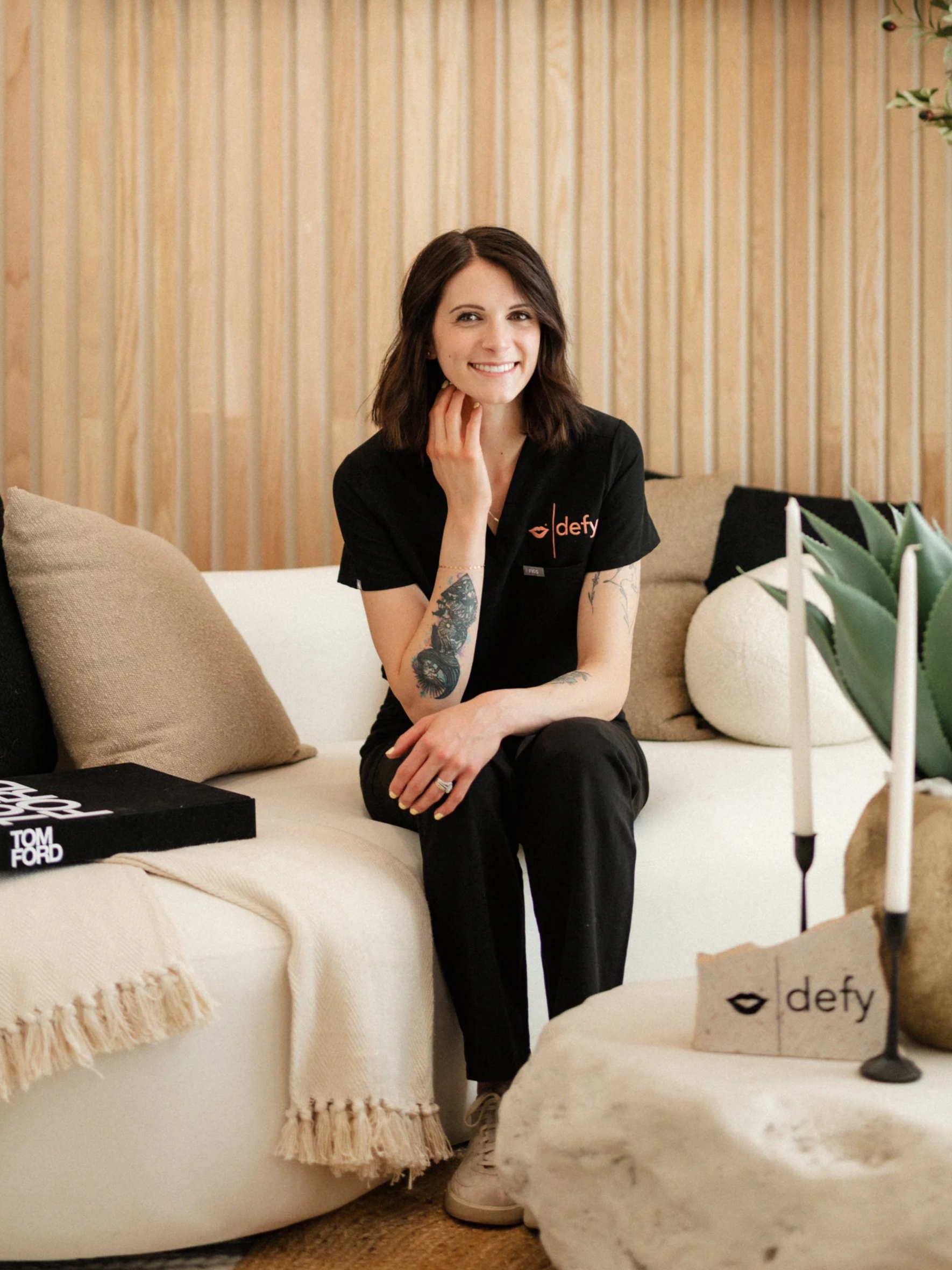 A woman with shoulder-length brown hair and tattoos on her arms, sitting on a beige couch, smiling, wearing a black shirt with the word 'defy' on it, in a cozy indoor space with plants and decorative pillows. Defy RS | Daxxify & Lip Filler in Pueblo