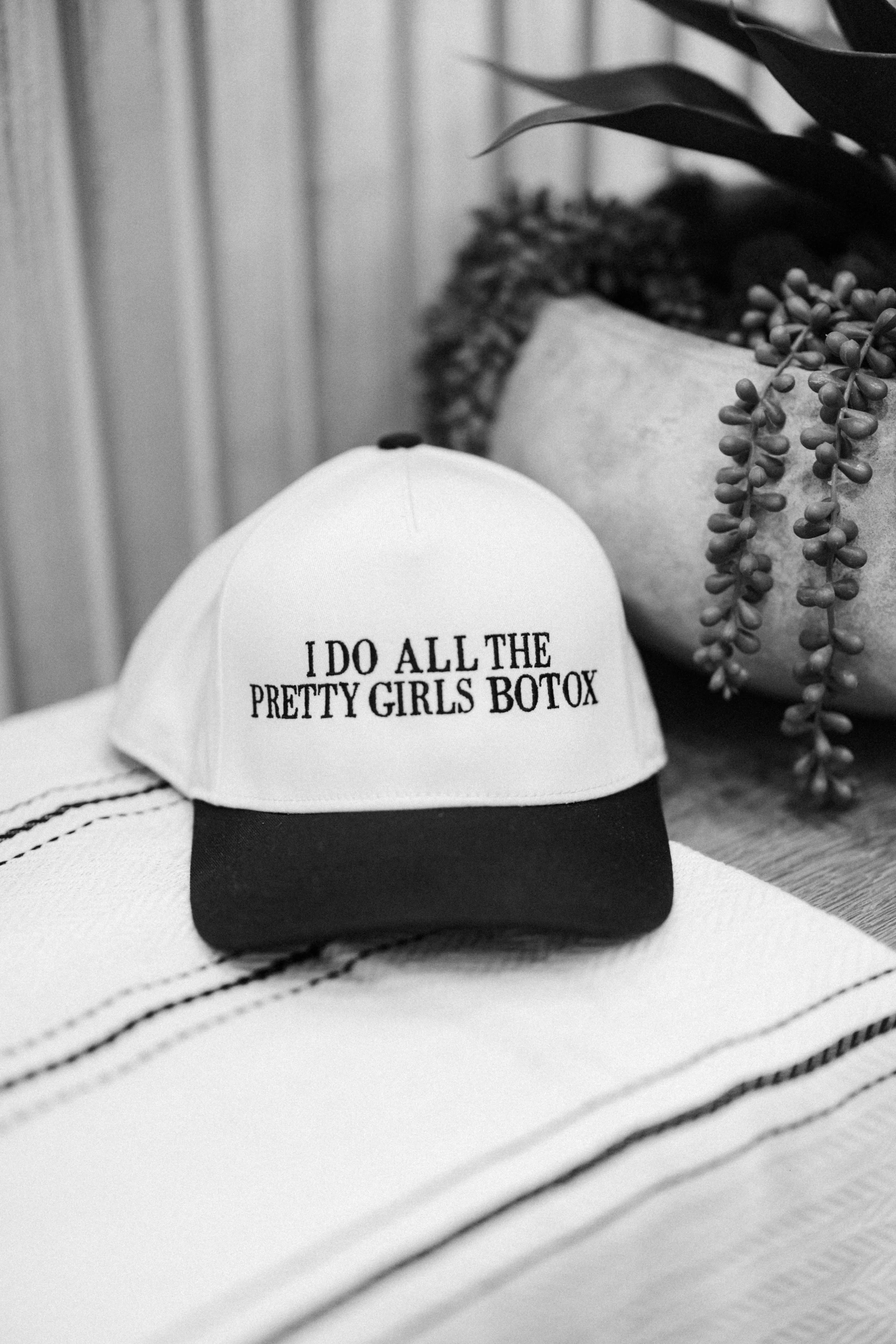 A white baseball cap with black brim and the words 'I DO ALL THE PRETTY GIRLS BOTOX' embroidered on the front, resting on a striped fabric surface with a potted plant in the background. Defy RS | Daxxify & Lip Filler in Pueblo West CO
