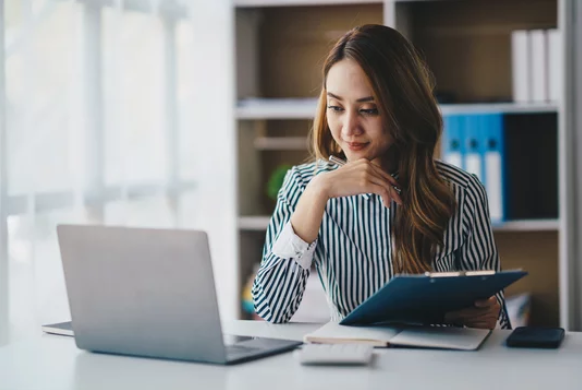 A woman sitting at a desk with an open laptop, reading a document, in a bright office setting.