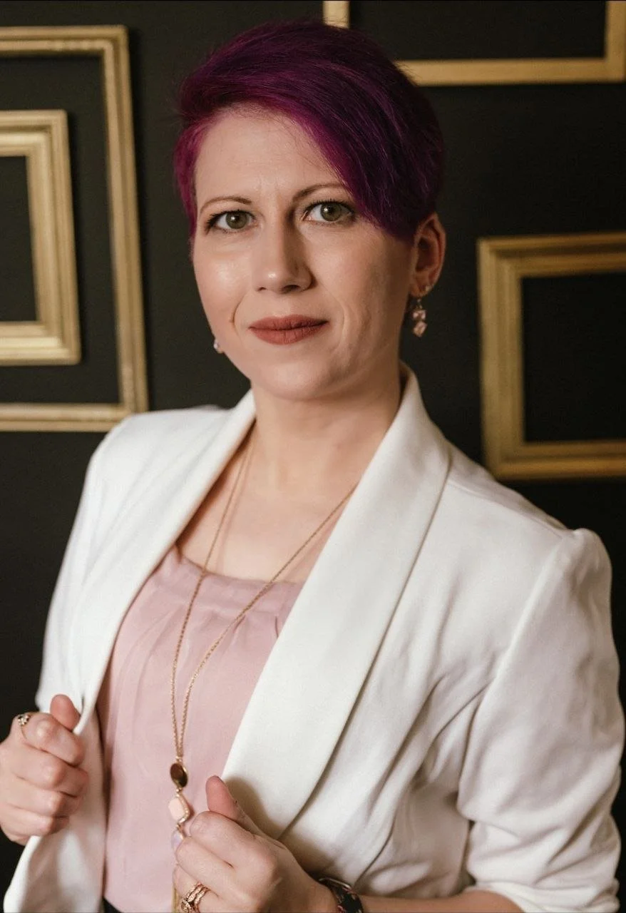 A woman with short, purple hair wearing a white blazer over a pink top, holding the edges of her blazer. She has earrings, a necklace, and rings, standing in front of a black wall with gold picture frames.
