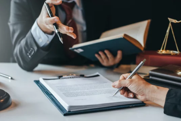Two people in business attire reviewing documents and reading a book in an office setting.