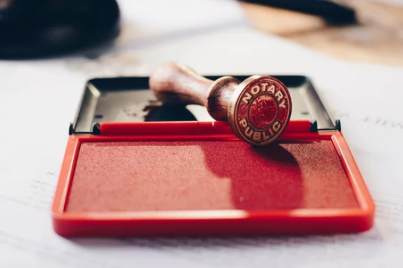 A notary public stamp resting on an open red stamp pad, with a blurred background of a document and office supplies.