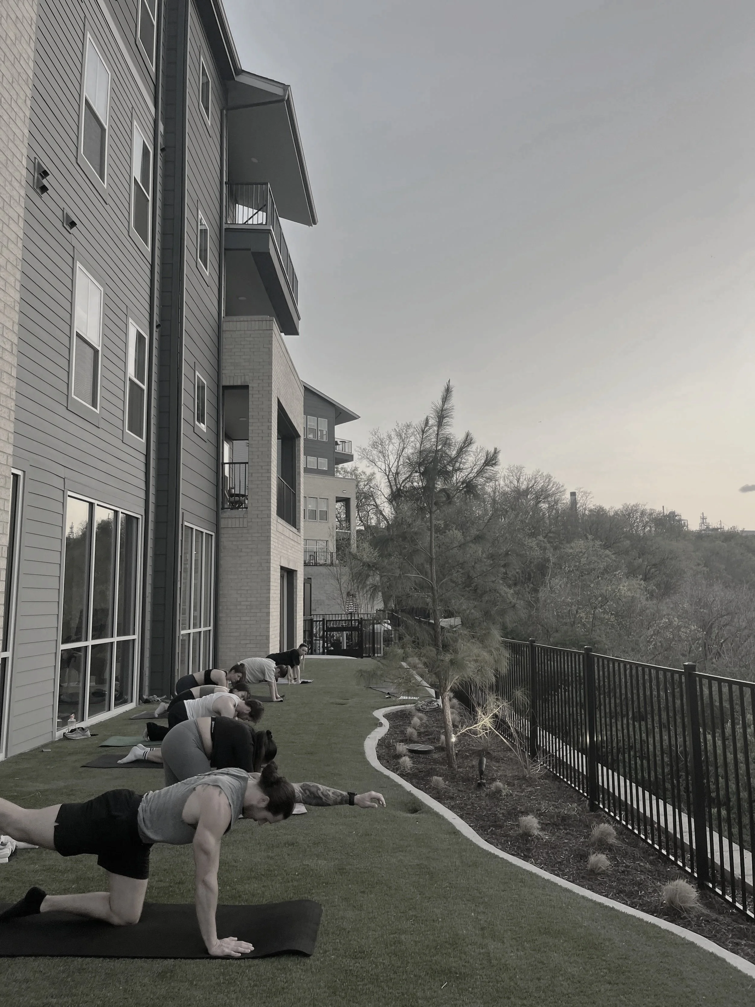 People practicing yoga outdoors on a balcony with a railing, overlooking trees and a gray sky.