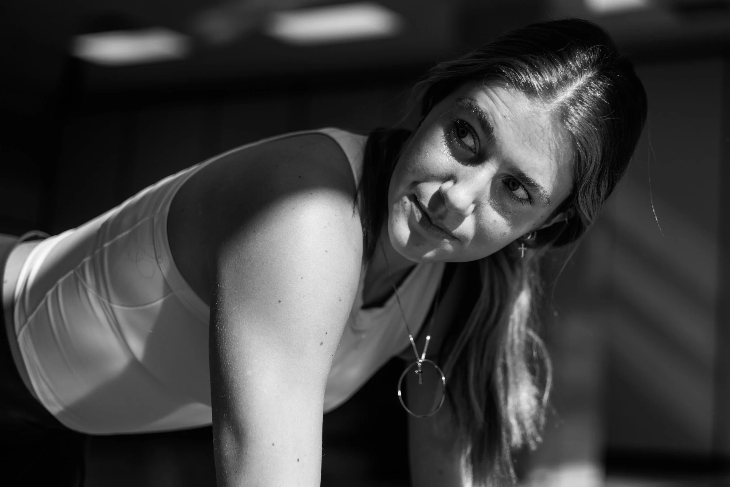 A woman with long hair, wearing a tank top and earrings, lounges indoors with sunlight casting shadows on her face.