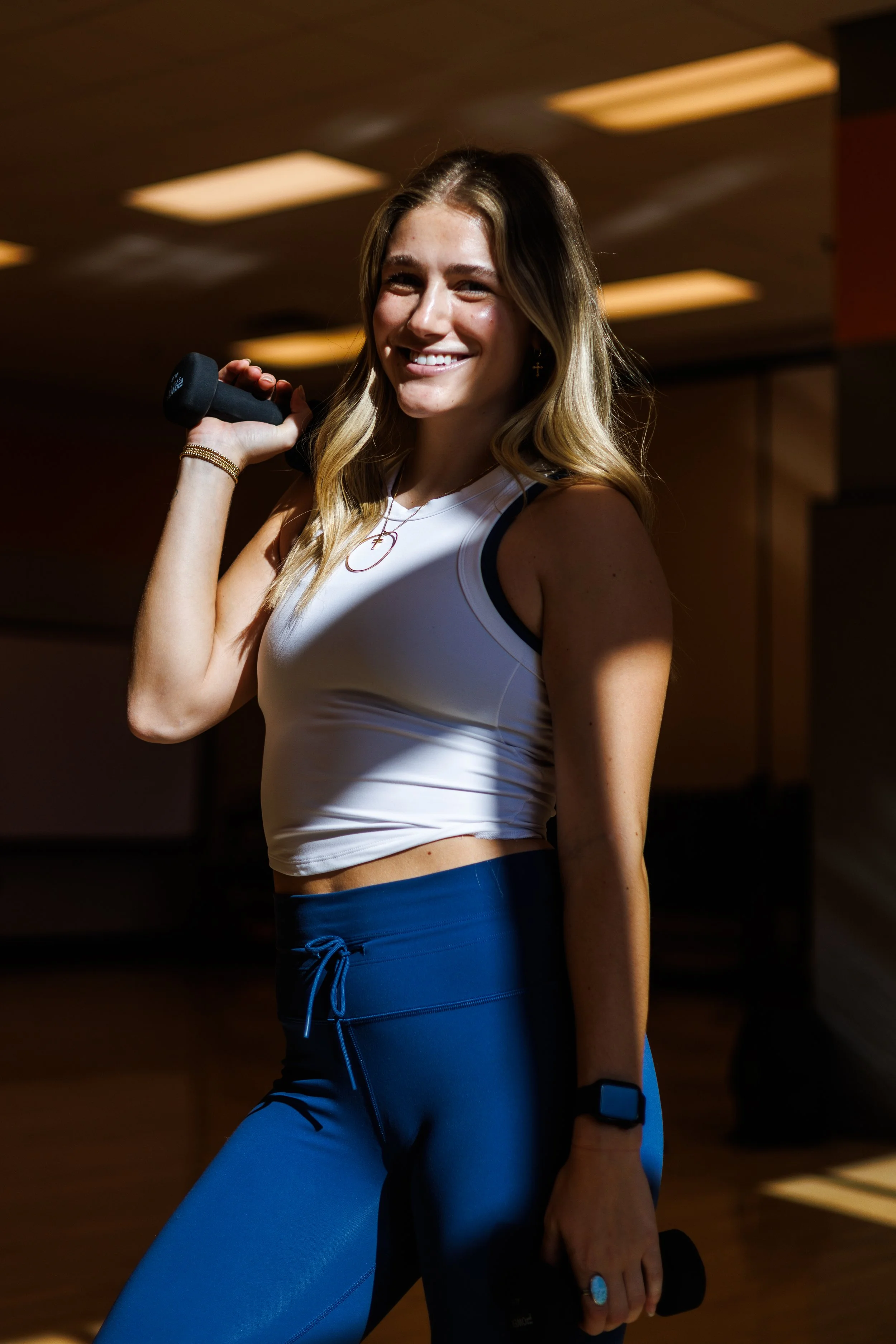 A young woman with long wavy hair smiling, holding a dumbbell on her shoulder and another dumbbell in her hand, wearing athletic clothing in an indoor fitness center.