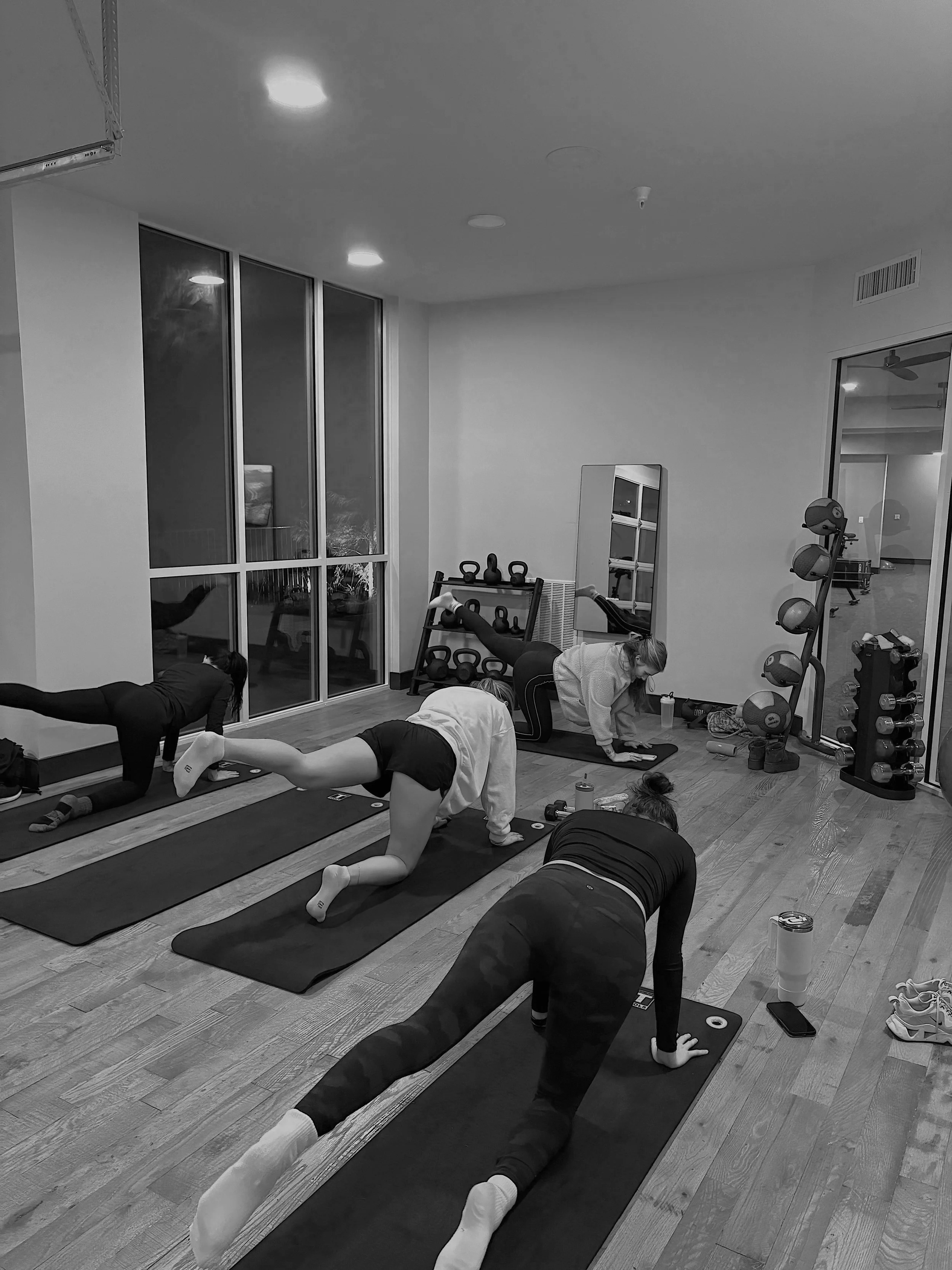 Four women practicing yoga on mats in a gym with kettlebells and weights in the background.