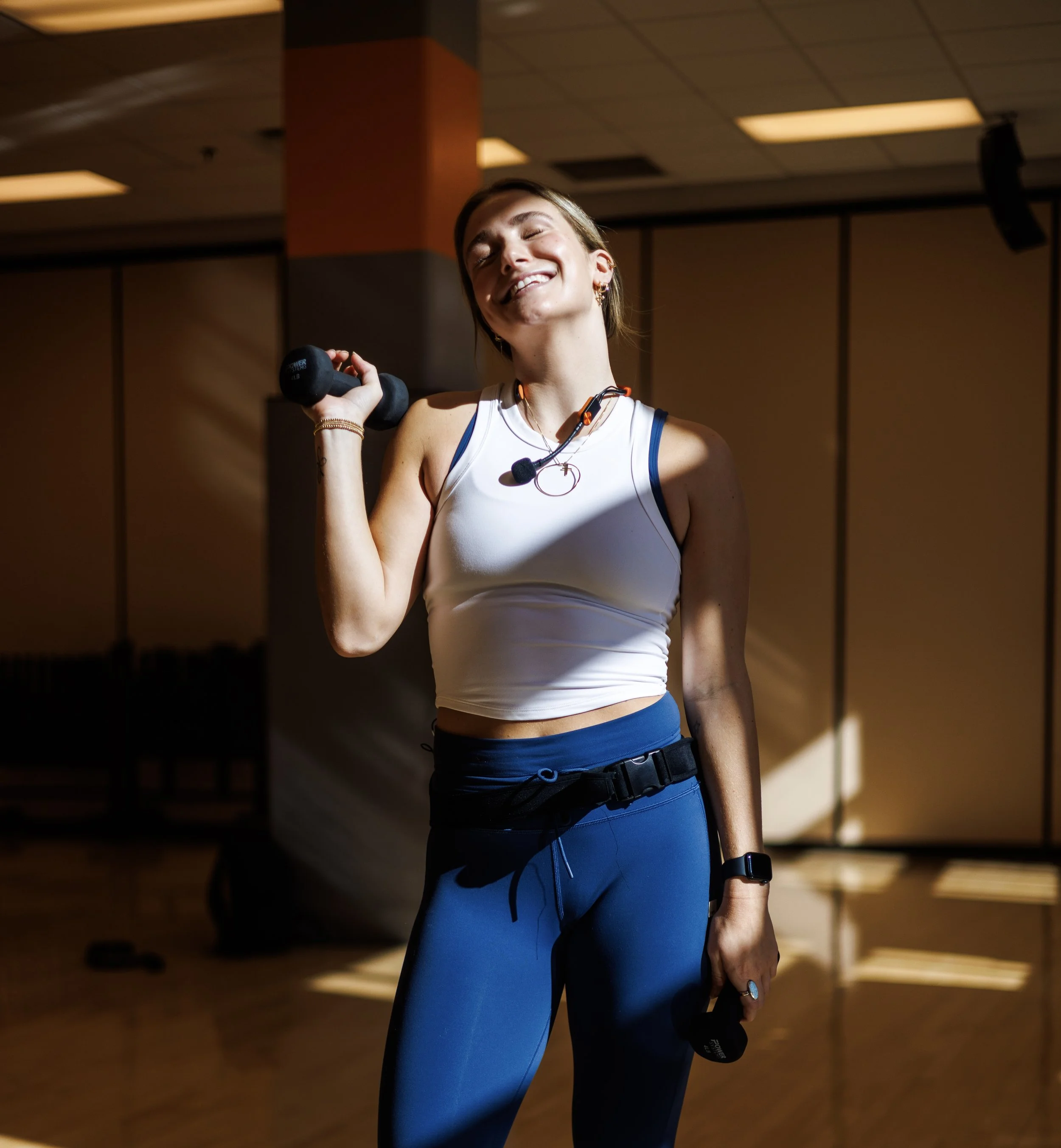 A woman smiling while holding dumbbells in a gym, wearing workout clothes and wireless earphones.