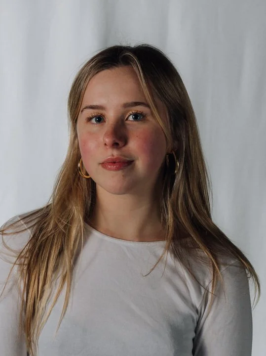 A young woman with long blonde hair, wearing a white top and gold hoop earrings, standing in front of a white background.