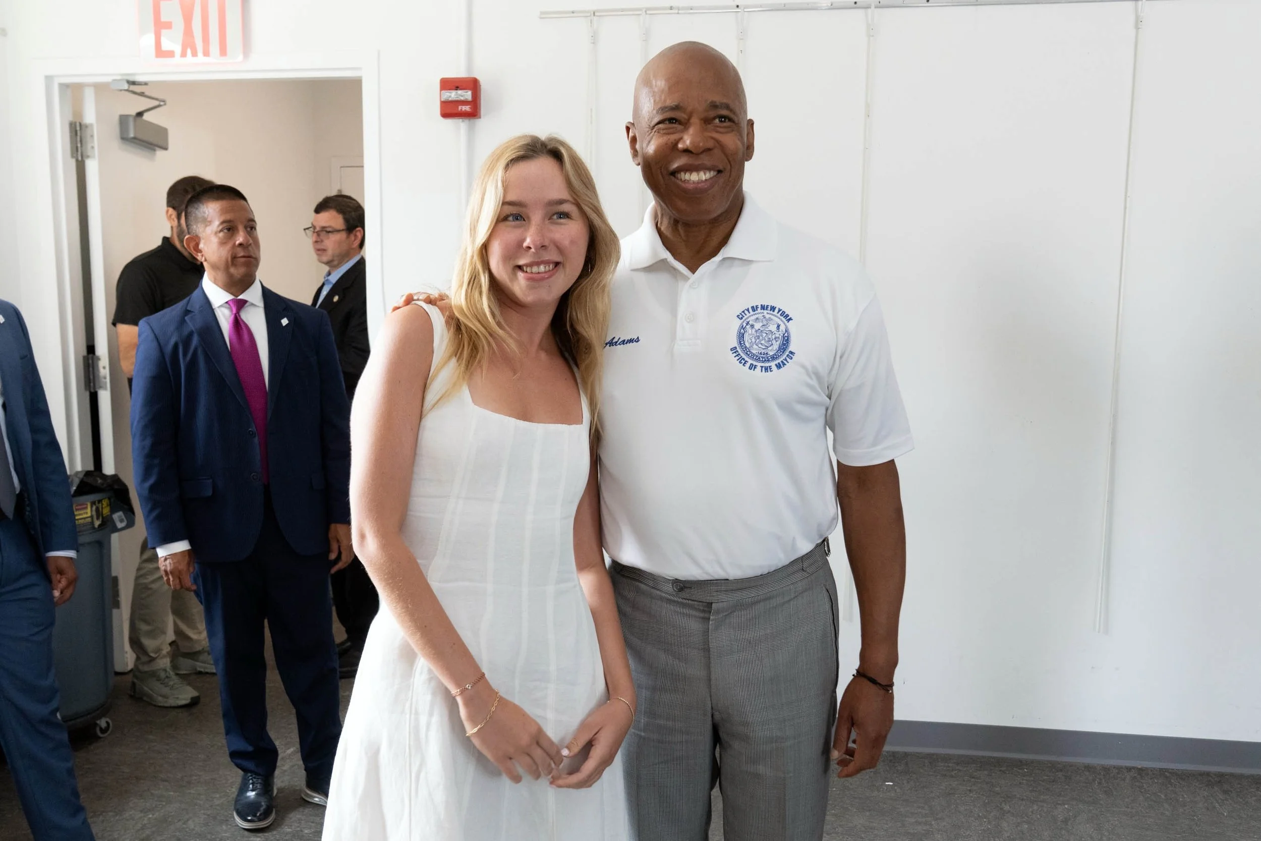 A woman and a man are smiling and standing side by side indoors, surrounded by other people, some of whom are wearing suits.