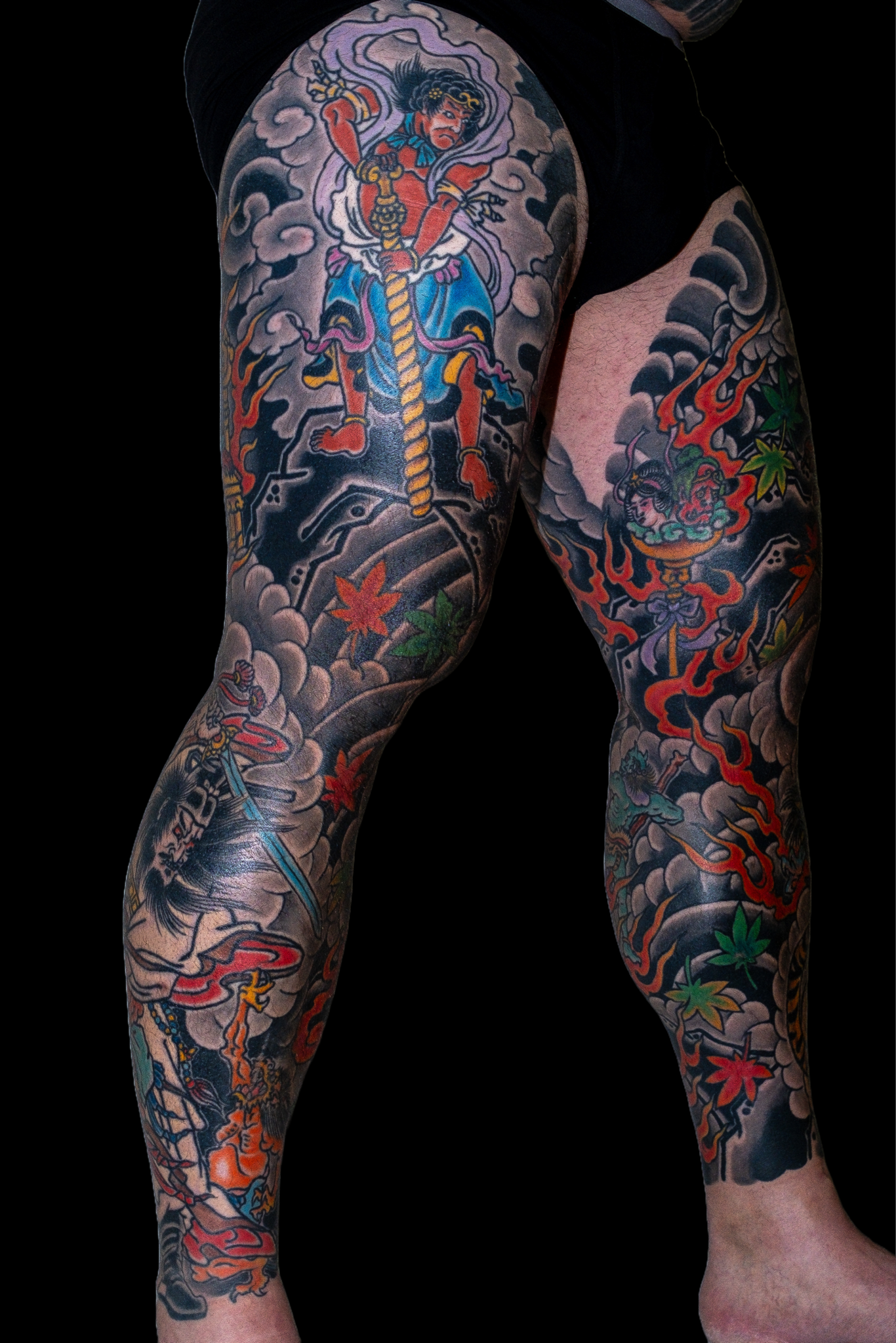 A person's legs and thigh with detailed traditional Japanese-style tattoos featuring a fierce warrior, clouds, flames, and maple leaves, with vibrant colors and intricate designs.