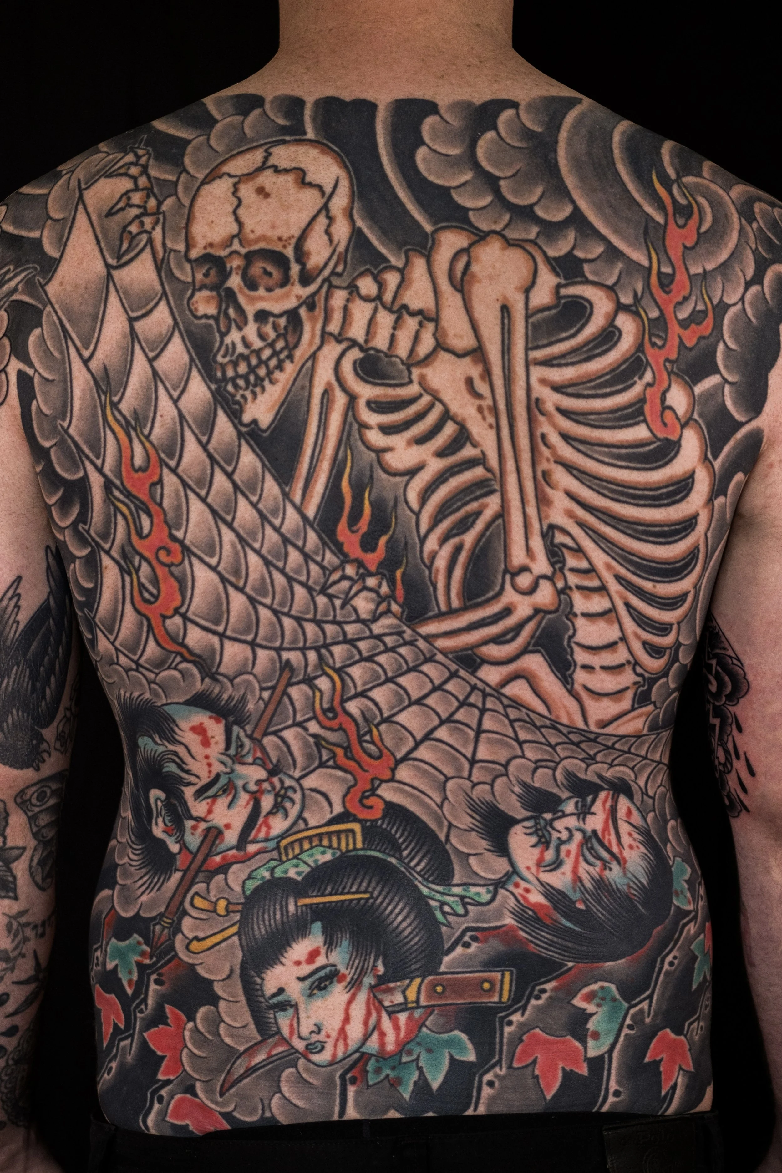 Tattoo of a skeleton with wings and flames on the chest and arms, and a Japanese woman with black hair and red lipstick on the abdomen.