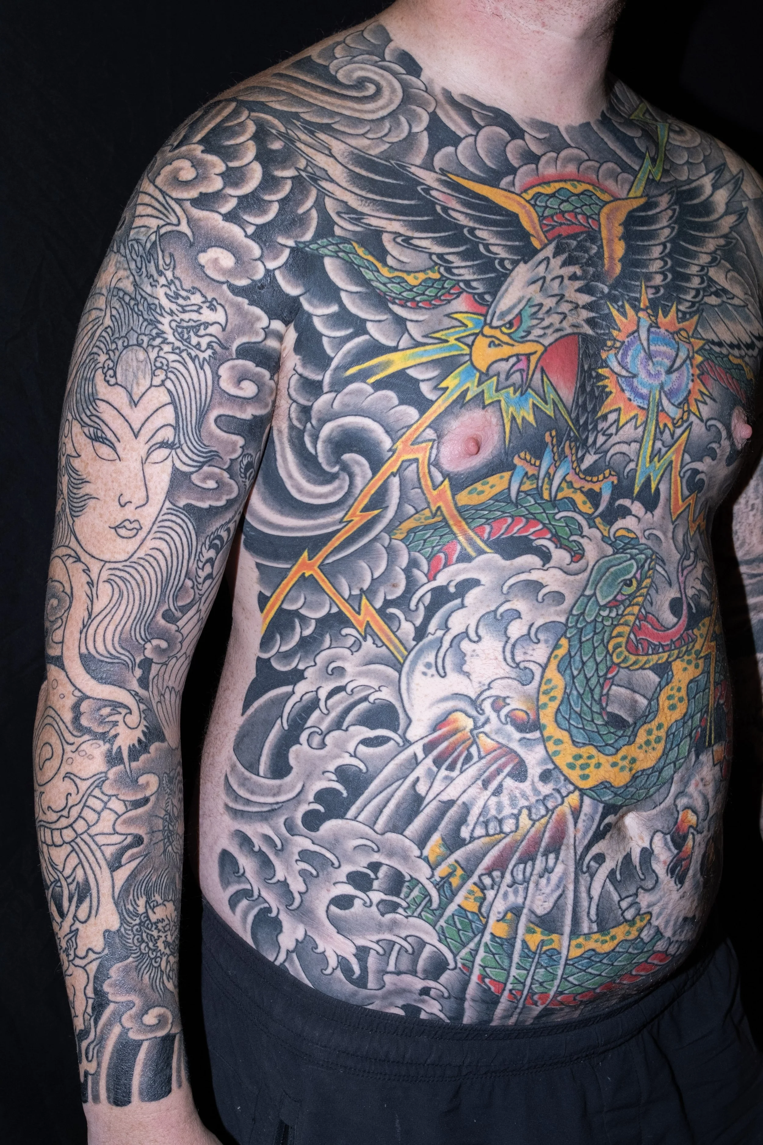 A man with extensive tattoos on his chest, arms, and shoulder, including a large eagle with spread wings and a snake, and a kimono-style face on his arm, all in black, gray, and colorful ink.