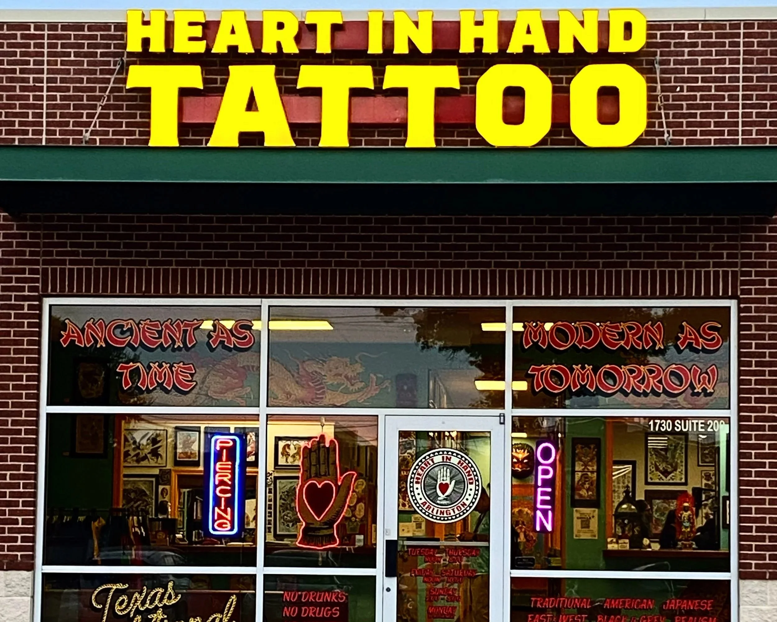 Sign on a storefront displaying yellow text that says 'Heart in Hand Tattoo' against a brick wall background.