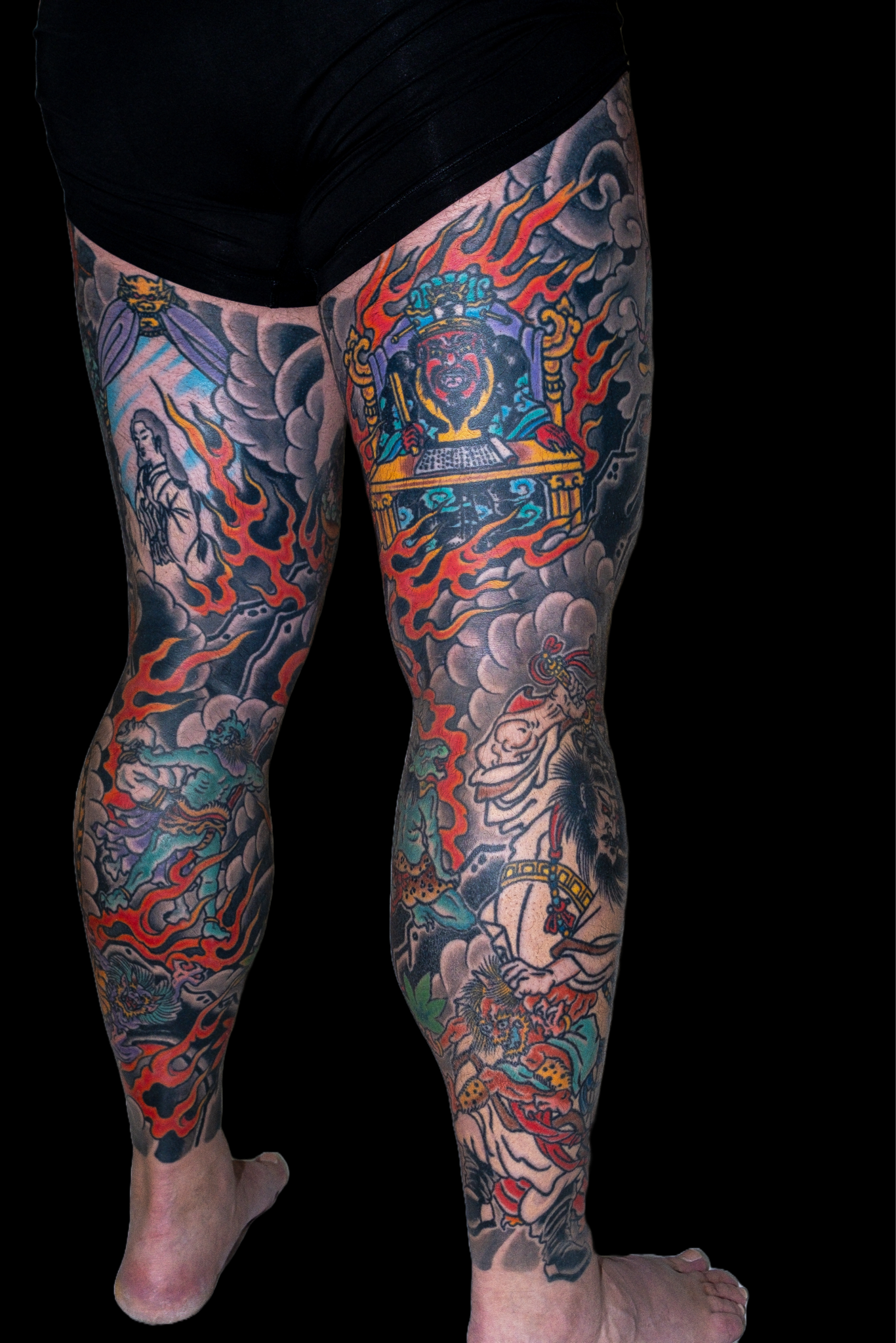 Close-up of a person's legs with traditional Japanese style tattoos featuring dragons, clouds, and a fierce warrior figure, against a black background.