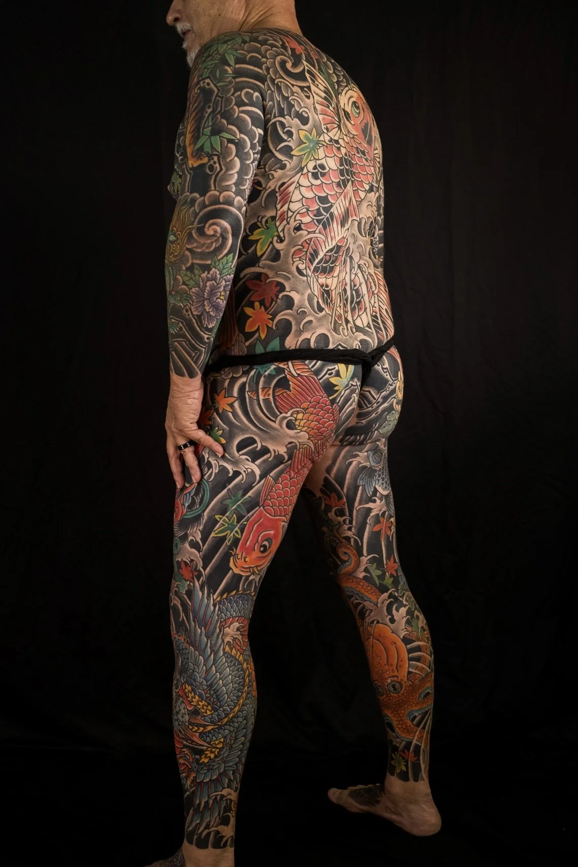 A man with extensive traditional Japanese style tattoos covering his body, wearing no clothes except a black waistband, standing against a dark background.