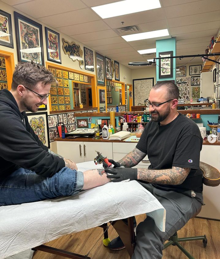 A man getting a tattoo on his leg at a tattoo shop. The tattoo artist is smiling and wearing black gloves. The shop is decorated with framed tattoo artwork on the walls.