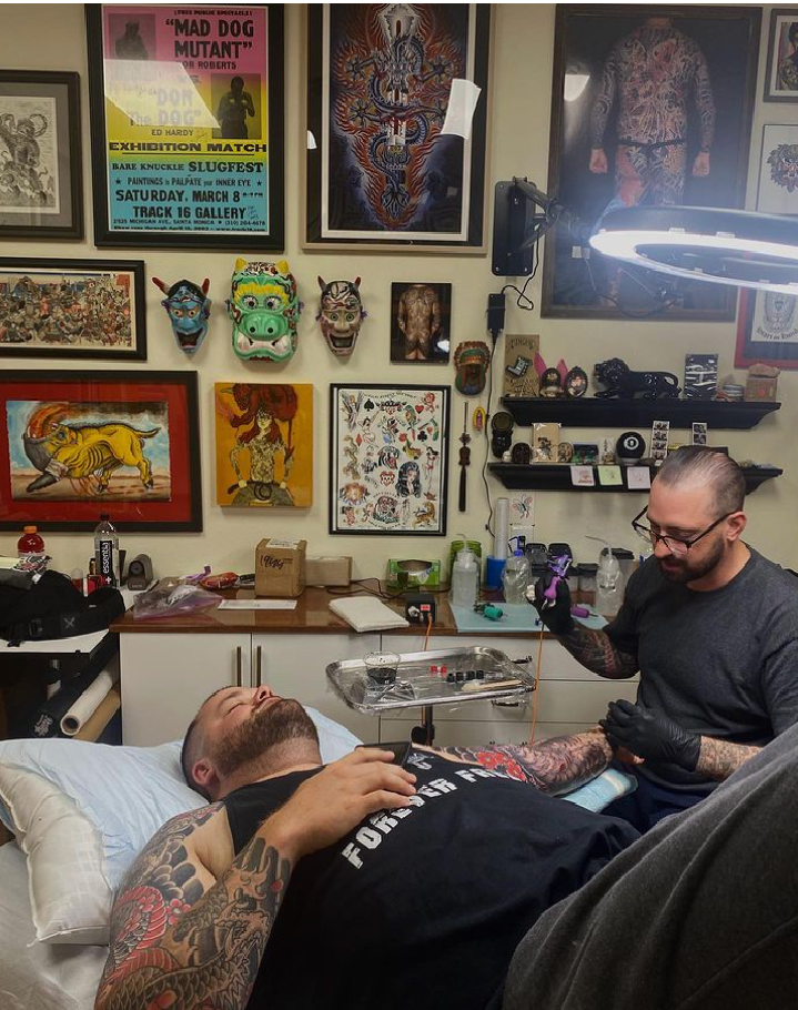 A man with tattoos lying on a hospital bed getting a tattoo from a tattoo artist in a tattoo studio decorated with colorful artwork and masks on the wall.