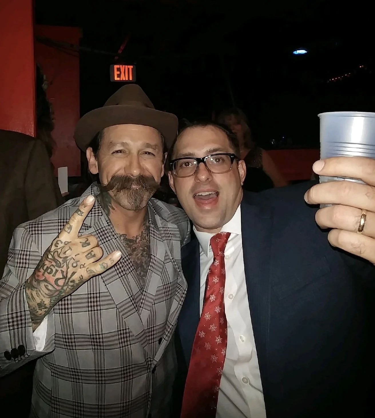Two men taking a selfie at a social event. The man on the left has a beard, tattoos, and is wearing a plaid suit, a hat, and making a hand gesture. The man on the right is wearing glasses, a suit, a tie, and is holding a cup, smiling with his mouth open.