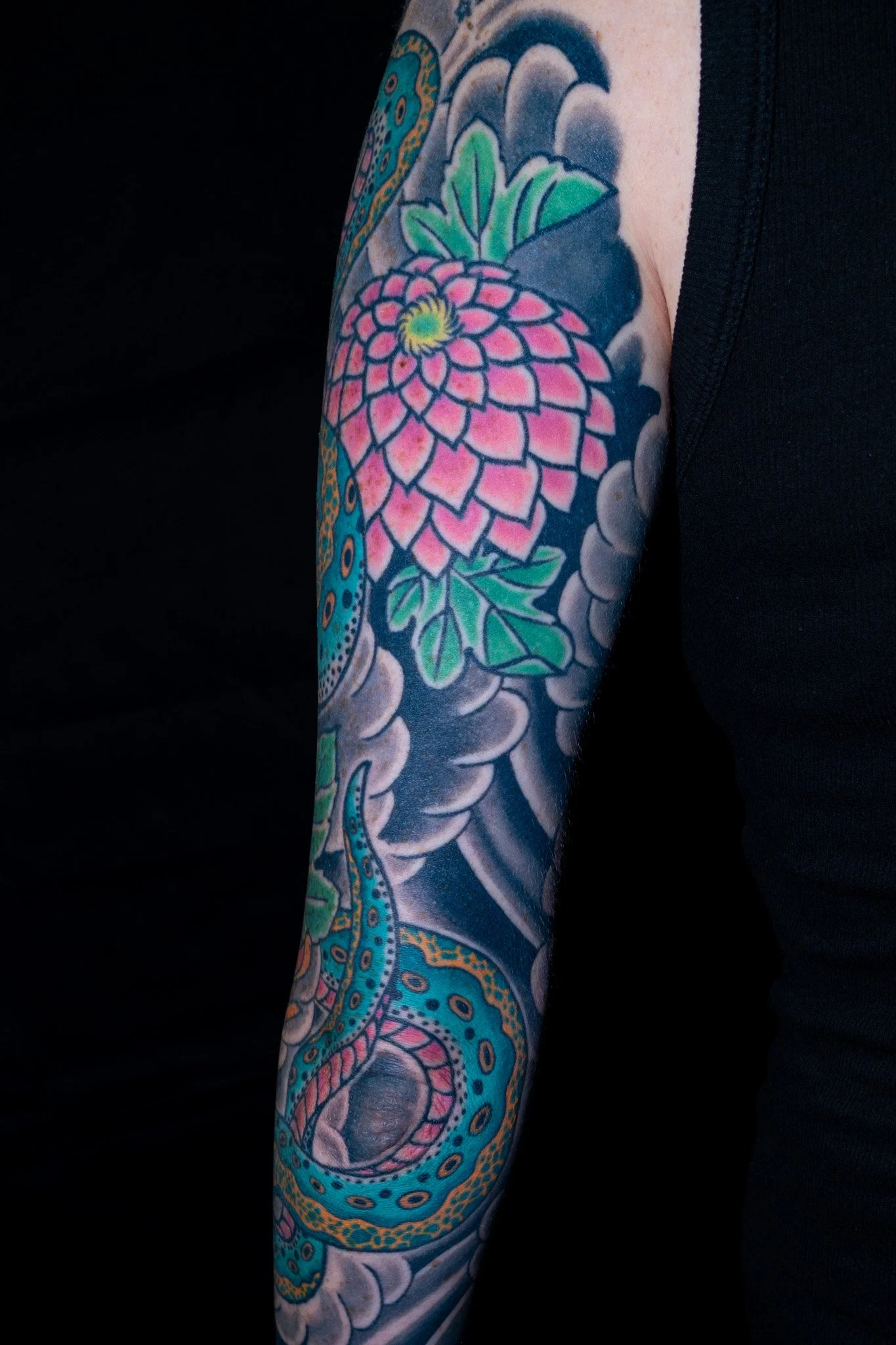 Colorful tattoo sleeve featuring a pink flower, green leaves, and a blue snake with intricate patterns on a person's arm. Tattoo by Joe Haasch. 