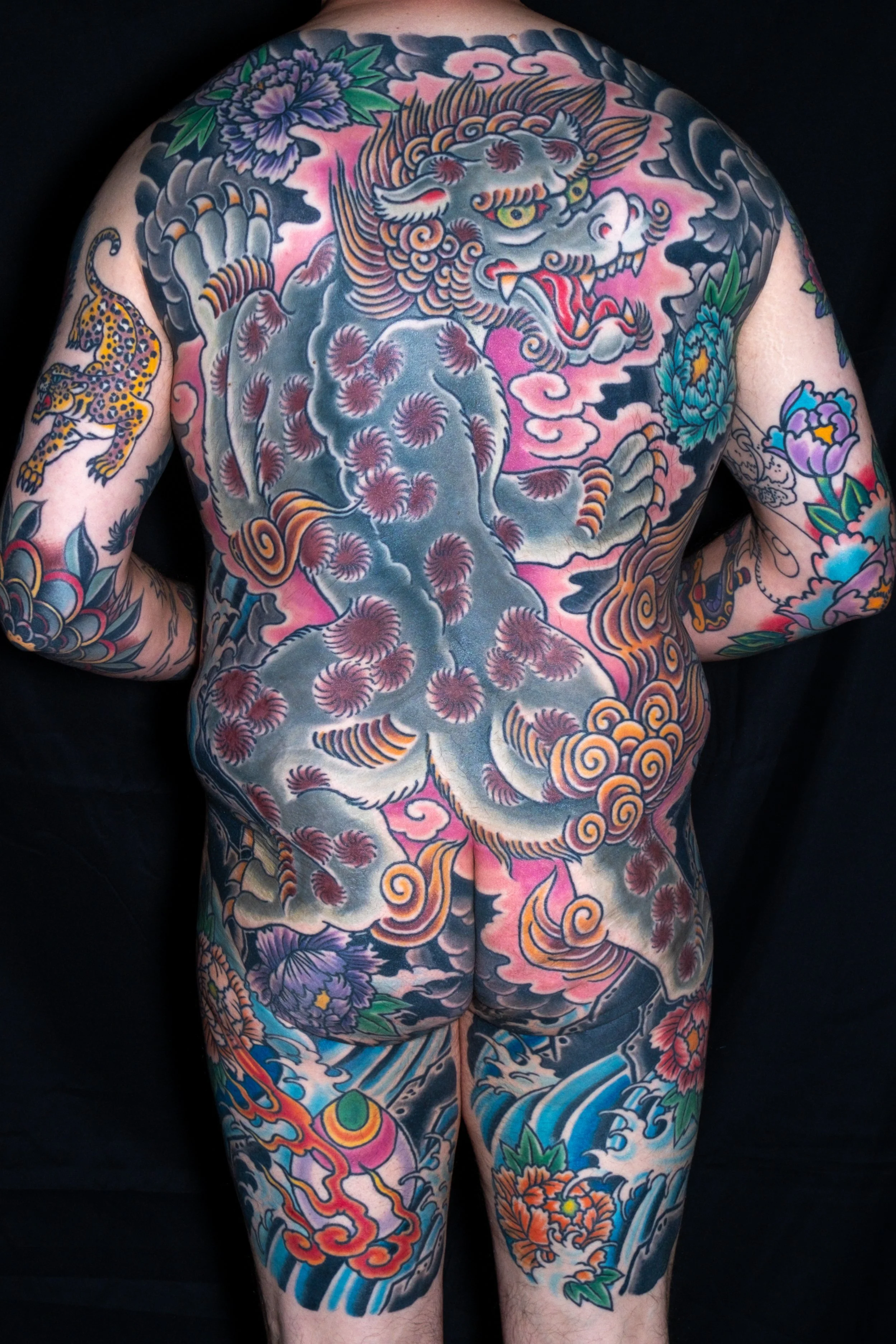 A person's back and legs covered in traditional Japanese style tattoos, featuring a large fearsome dragon on the back, surrounded by clouds, flowers, and swirling patterns.