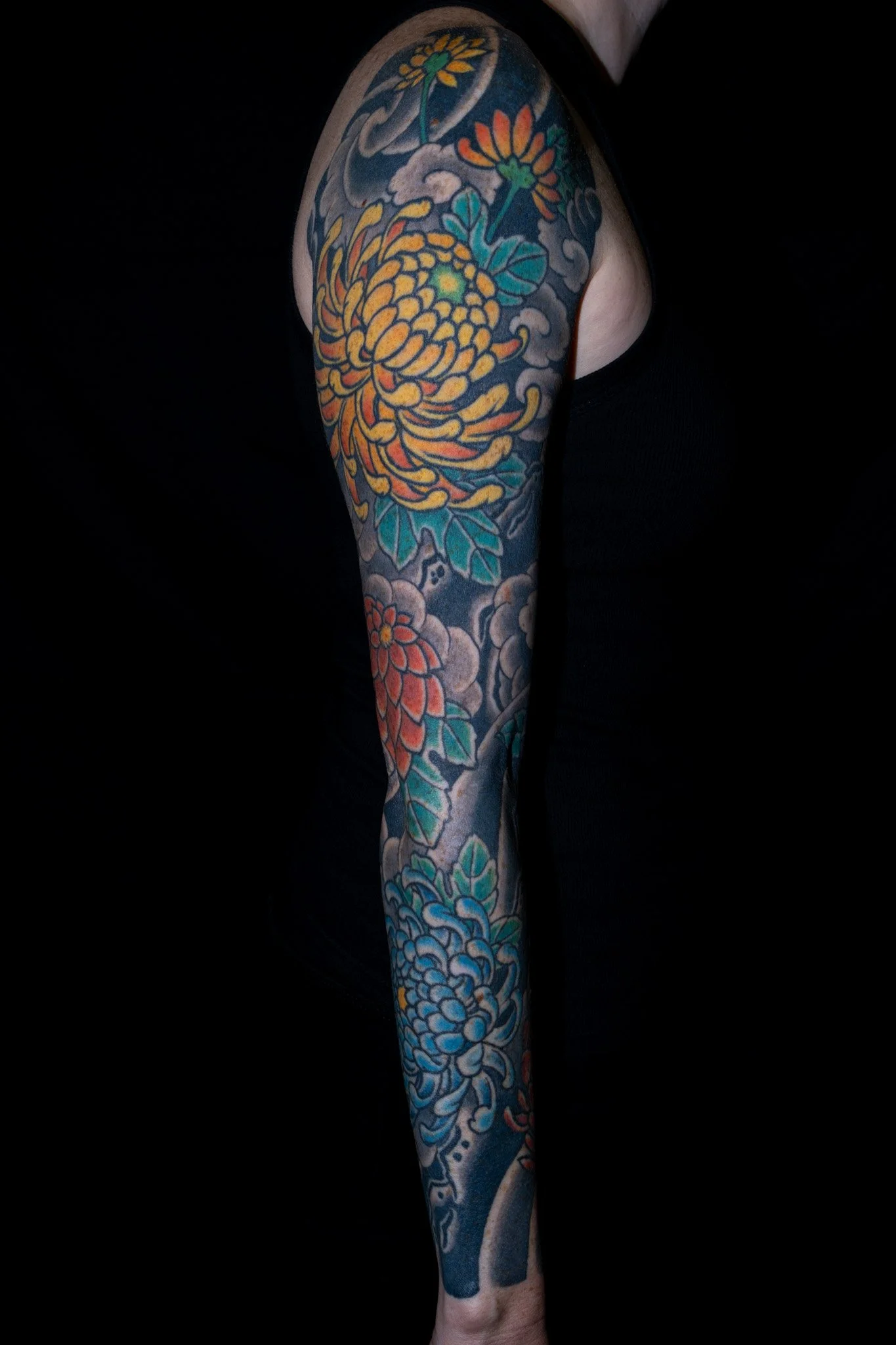 A person's arm and shoulder tattoo featuring colorful flowers, clouds, and intricate patterns on a dark background. Tattoo by Joe Haasch. 
