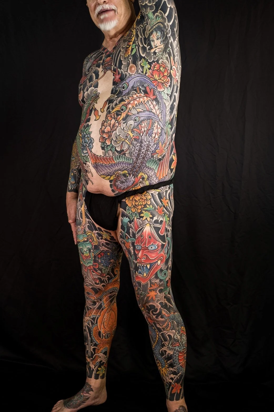 A man with a white beard and mustache, partially visible, standing against a black background. He is shirtless and wearing black underwear, showcasing extensive colorful Japanese-style tattoos covering his arm, chest, torso, and legs, including drago