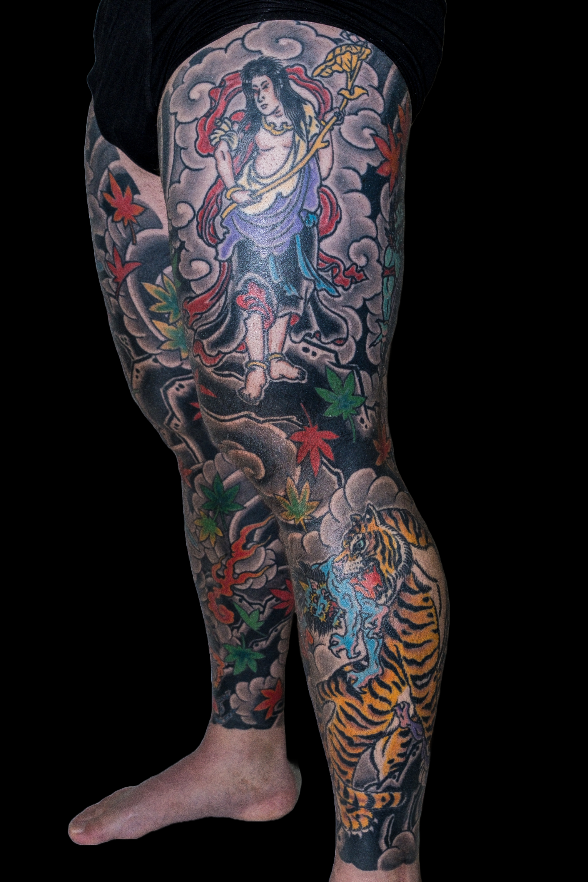 Tattoo of a woman with long hair holding a staff, surrounded by clouds and maple leaves, on a person's leg.