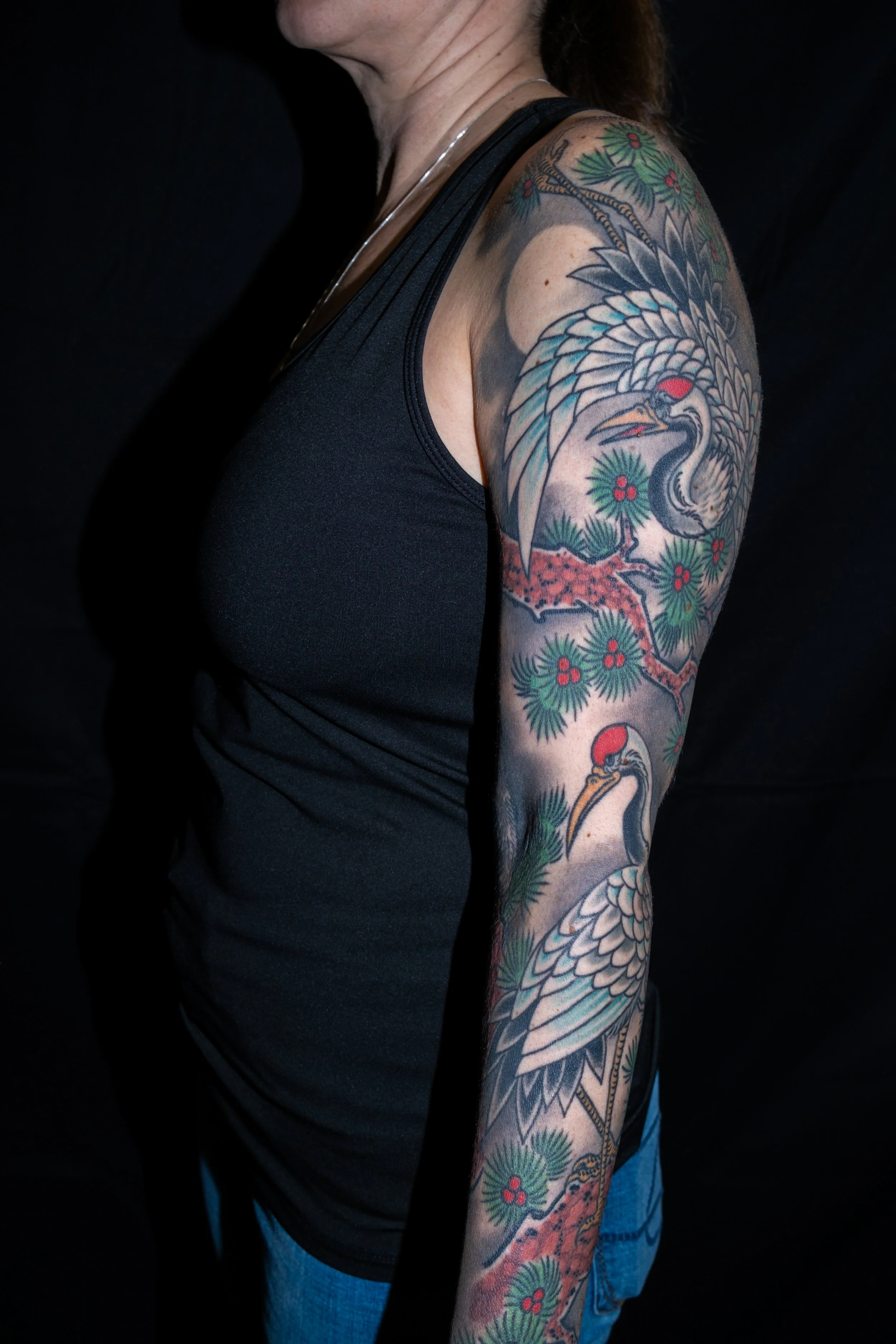 Tattooed woman wearing a black sleeveless top and blue jeans, showcasing a detailed tattoo sleeve featuring a crane, pine branches, and red berries on her upper arm. Tattoo by Joe Haasch. 