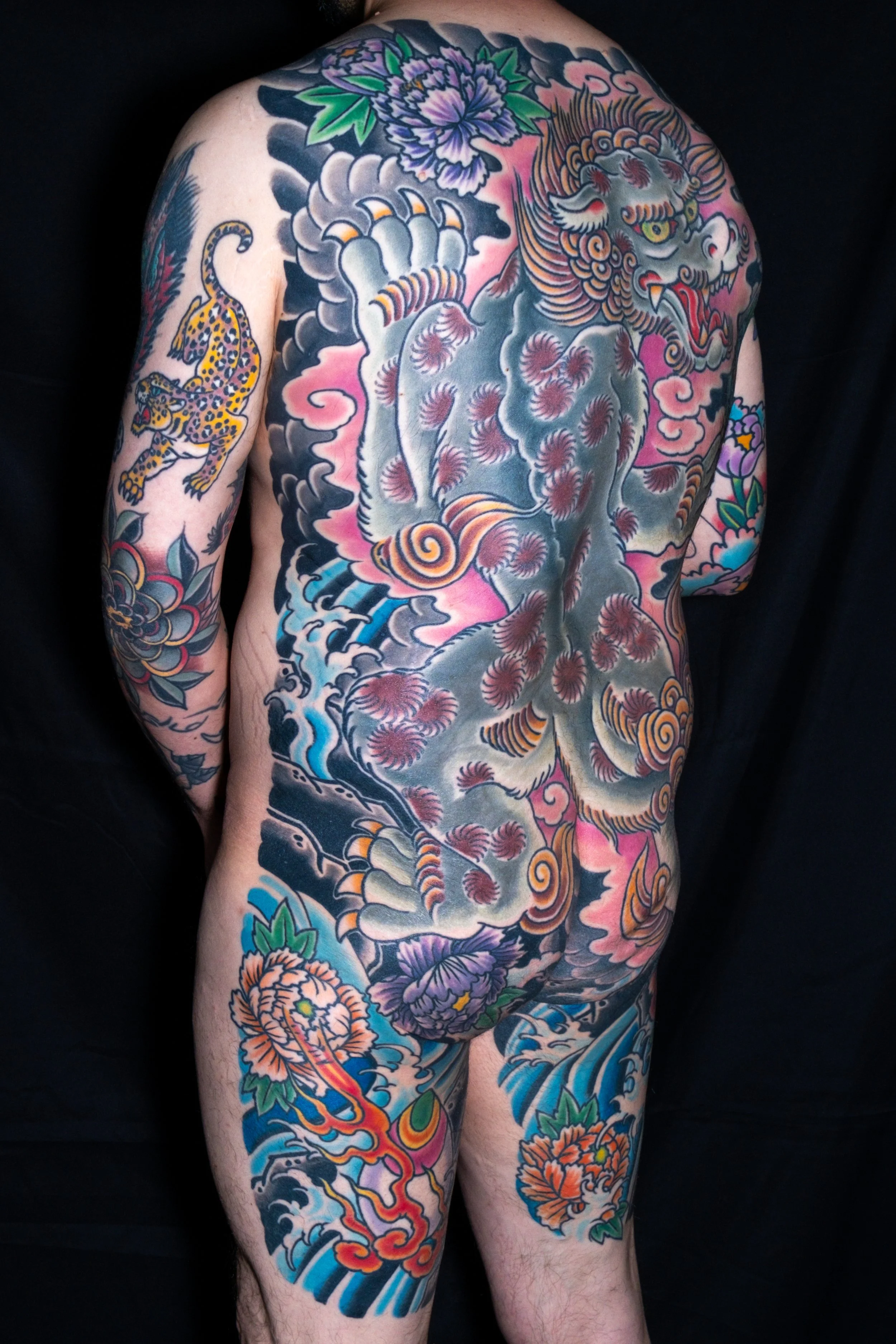 Full back tattoo of a fierce lion with a mane, surrounded by colorful flowers, clouds, and waves in traditional Japanese style.