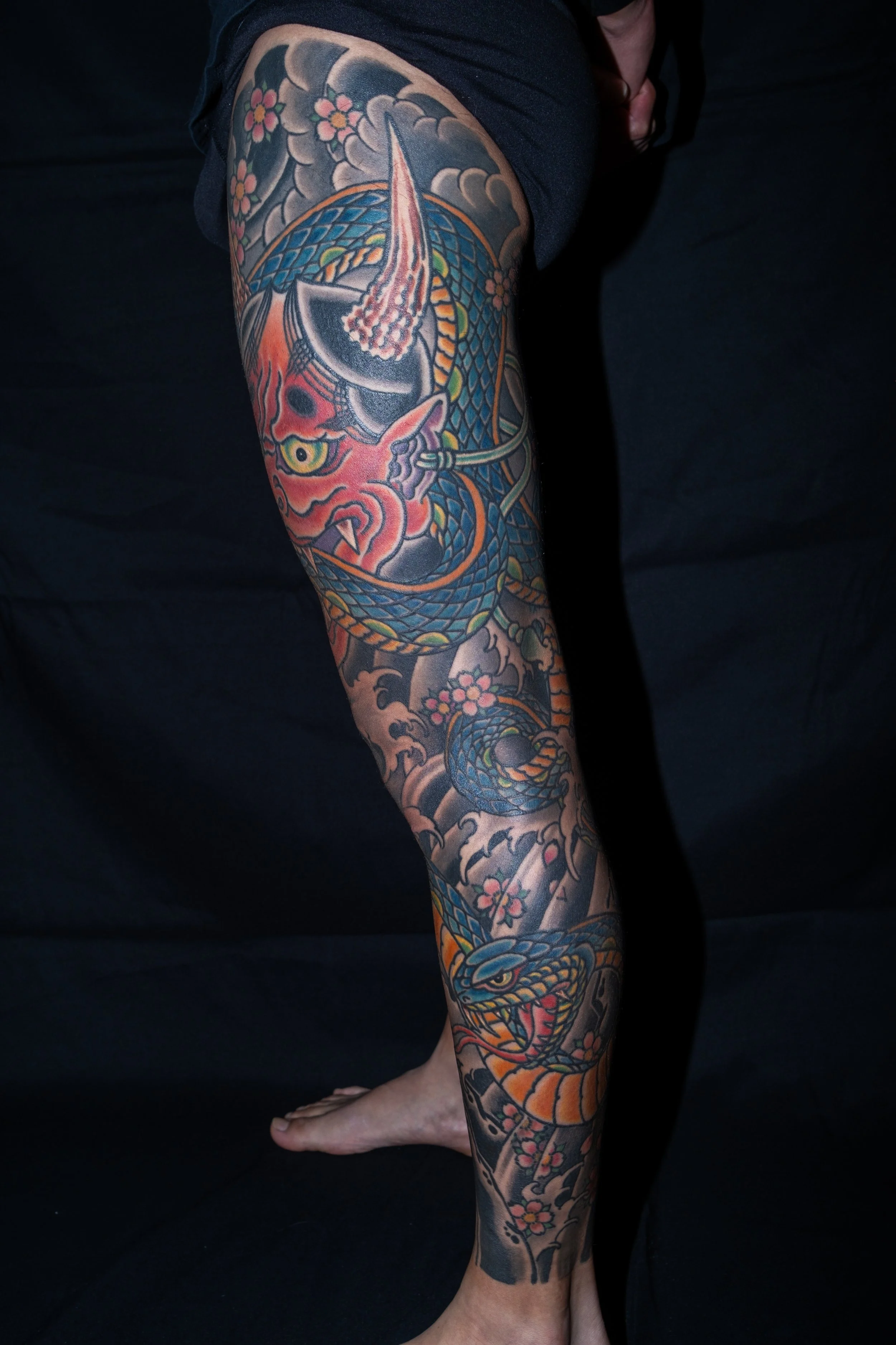 A person's arm with a detailed colorful tattoo featuring a snake intertwined with cherry blossoms, clouds, and other traditional Japanese-style design elements. Tattoo by Joe Haasch. 