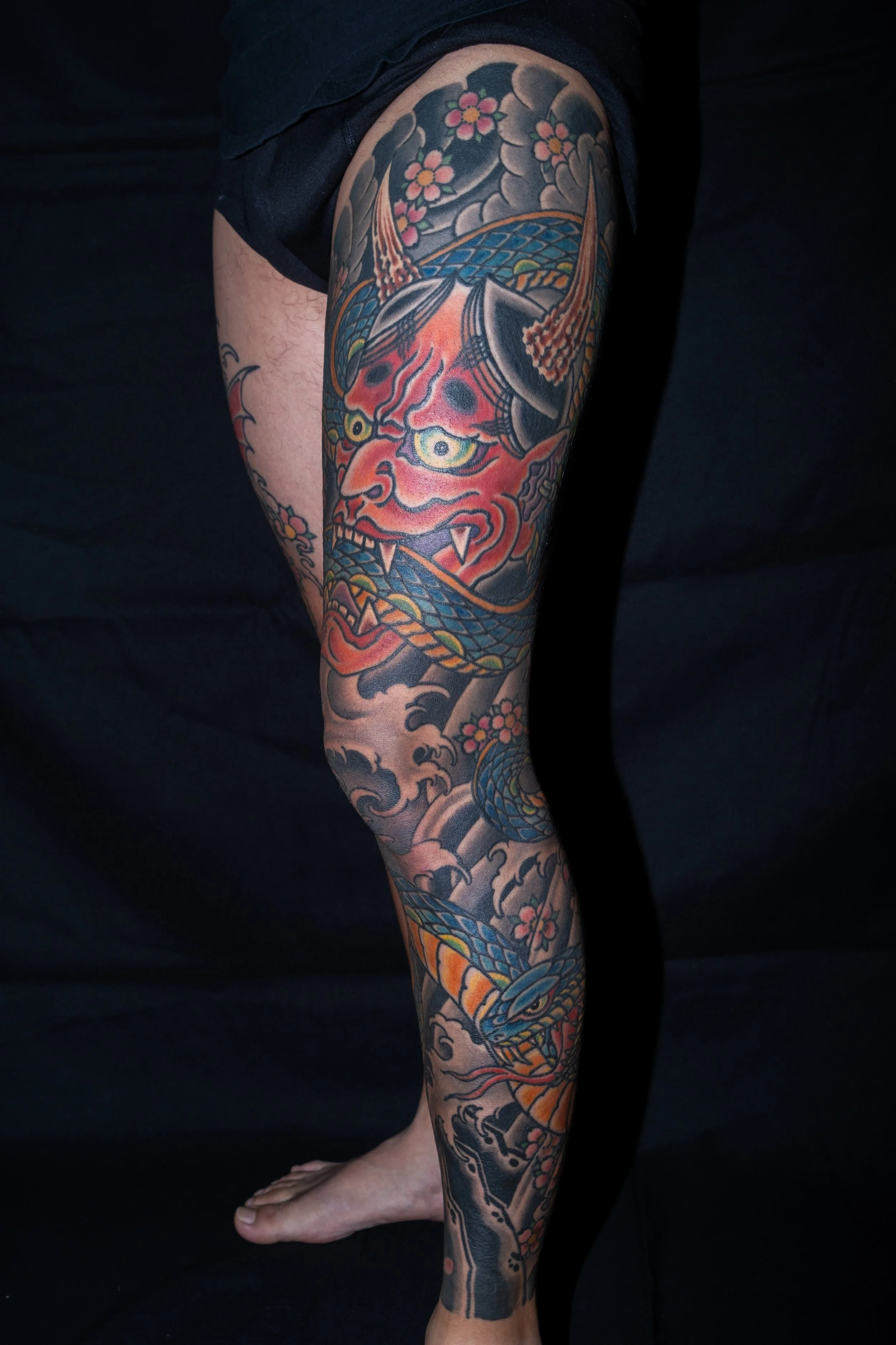 Colorful Japanese-style tattoo featuring a fierce demon face with horns and a dragon intertwined with cherry blossoms on a person's leg. Tattoo by Joe Haasch. 