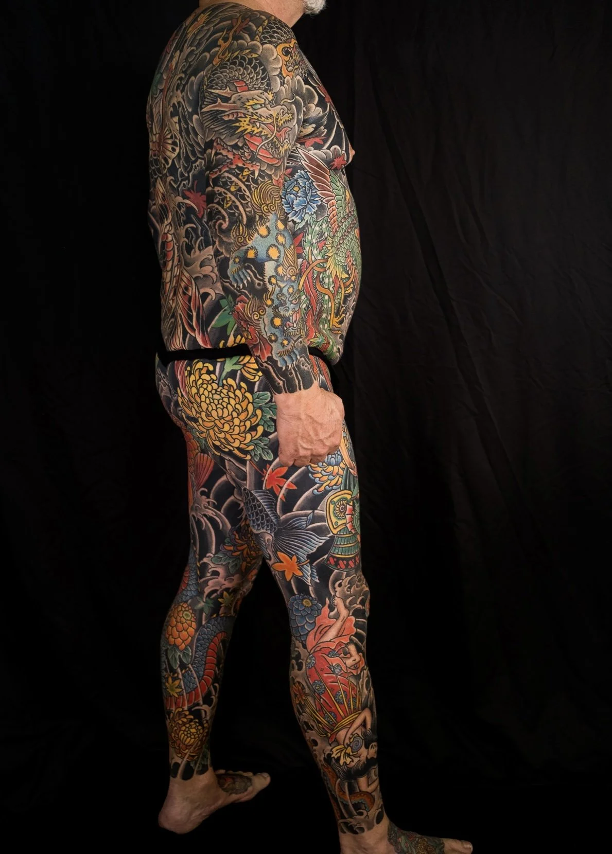 A man with extensive Japanese-style full-body tattoos, including dragons, flowers, and traditional motifs, standing barefoot against a black background.