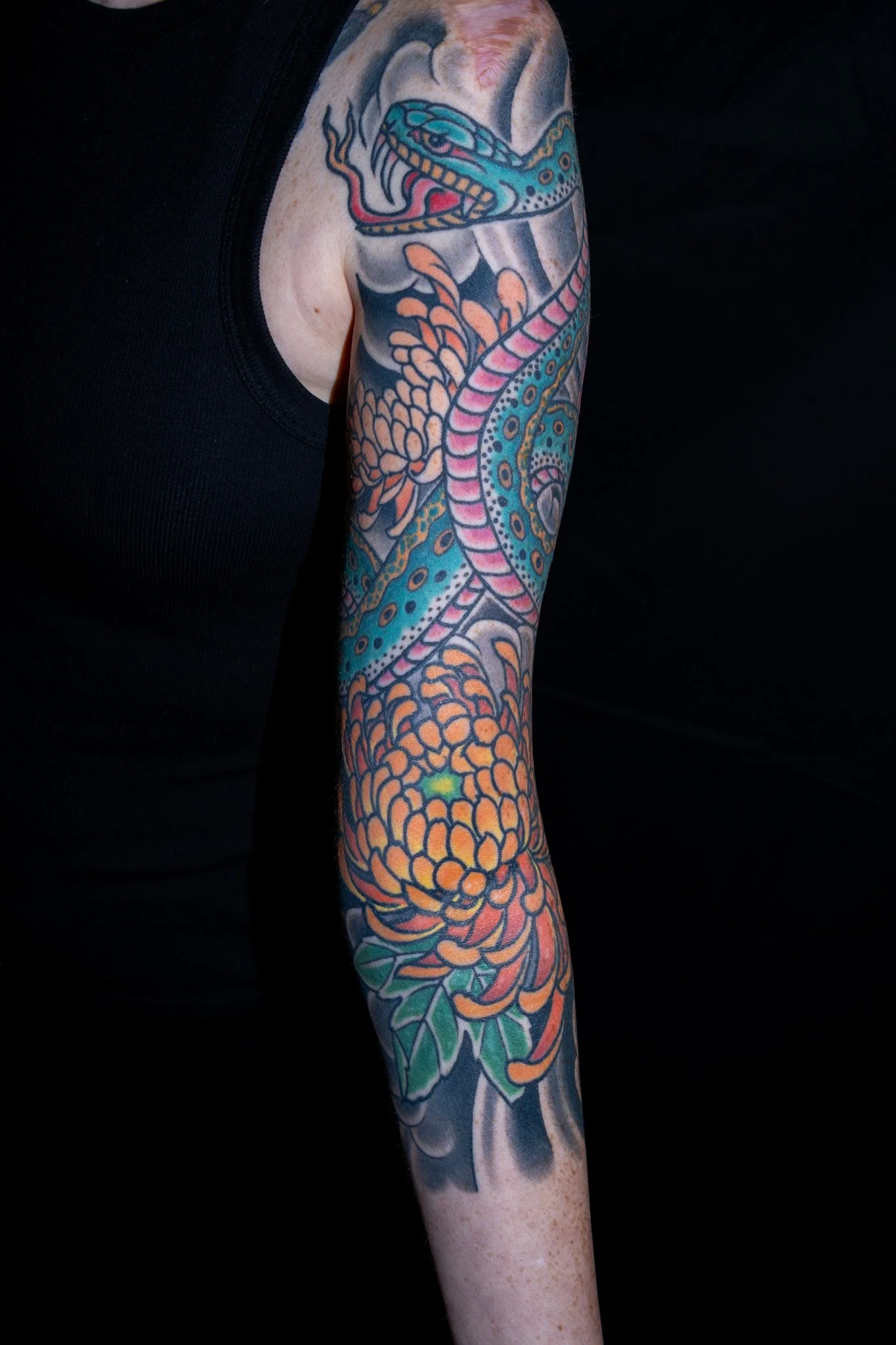 Colorful tattoo of a snake with vibrant scales wrapping around a person's arm, set against a black background. Tattoo by Joe Haasch. 