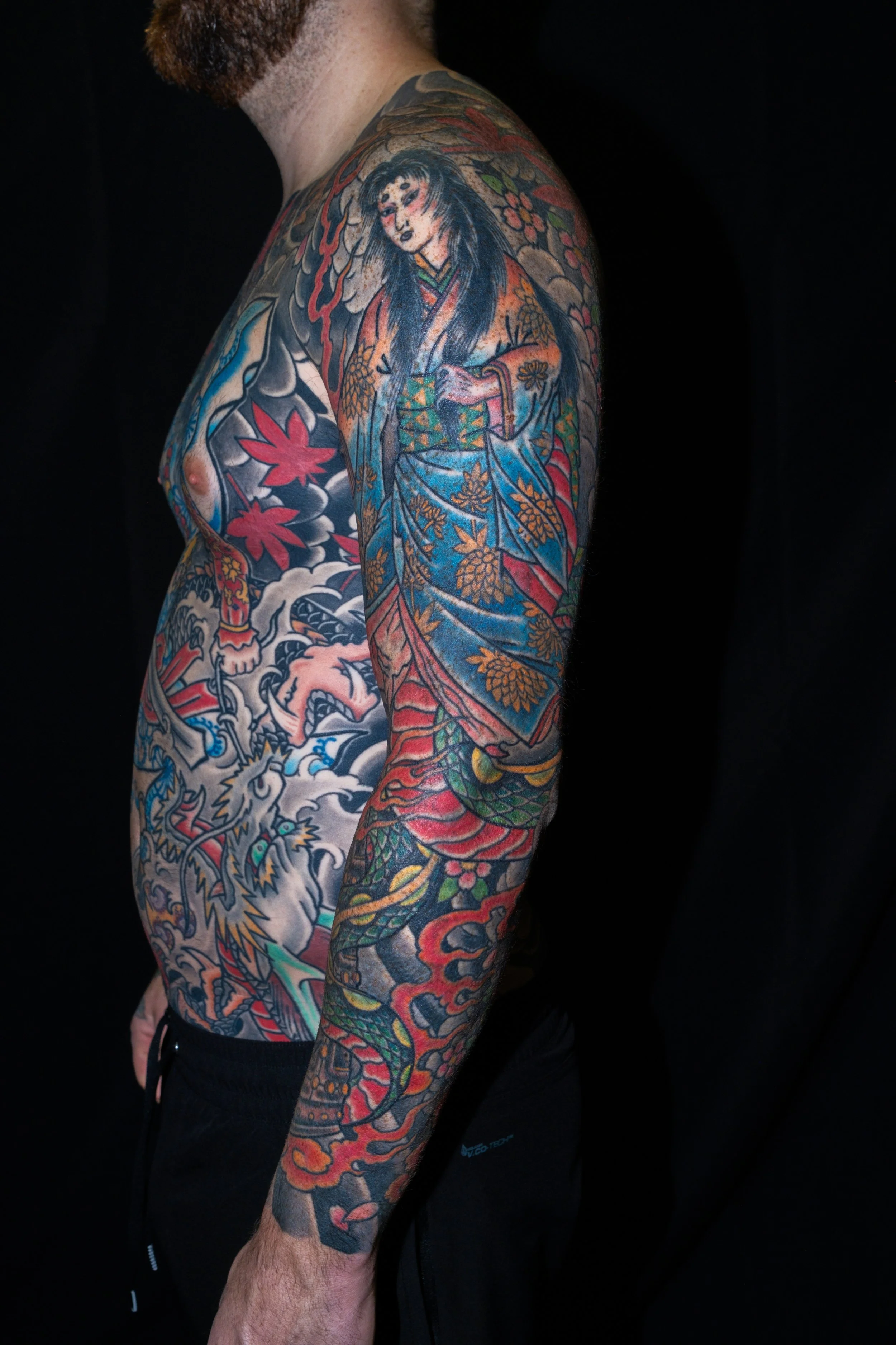 A person with a full sleeve tattoo of traditional Japanese art, including a woman, a dragon, floral patterns, and clouds, against a black background.
