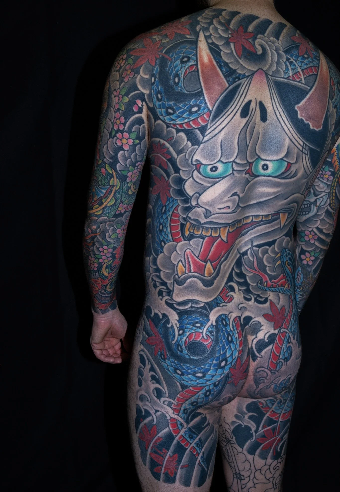 A person with extensive traditional Japanese style tattoos covering their back, shoulder, arm, and leg. The tattoo features a large, fierce dragon intertwined with clouds and red maple leaves, with vivid colors including blue, red, and black.