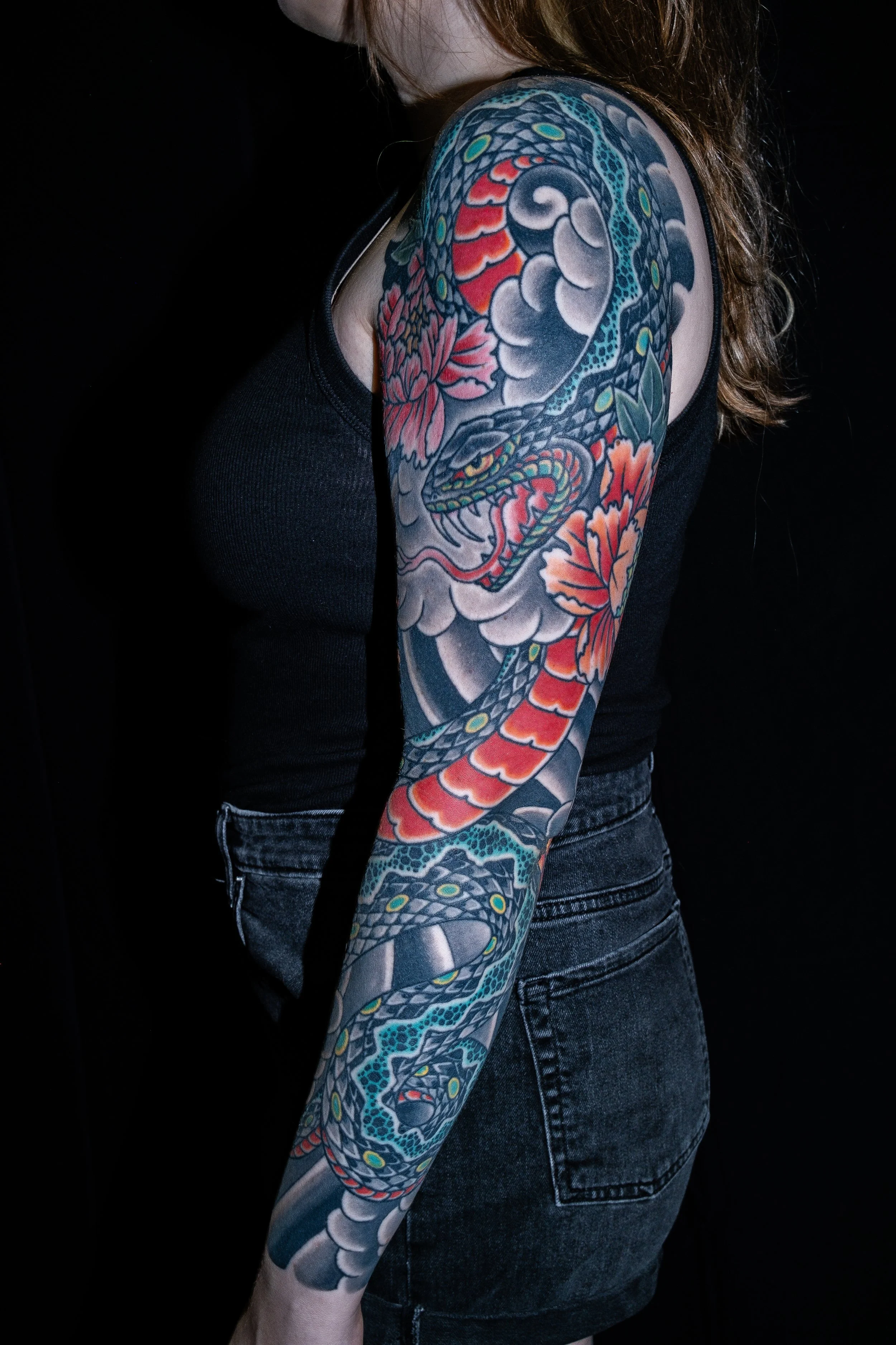A person with long hair and a visible face turned away, wearing a black top and dark jeans, displaying a colorful traditional Japanese-style tattoo sleeve of a snake and flowers on their arm, against a black background. Tattoo by Joe Haasch. 