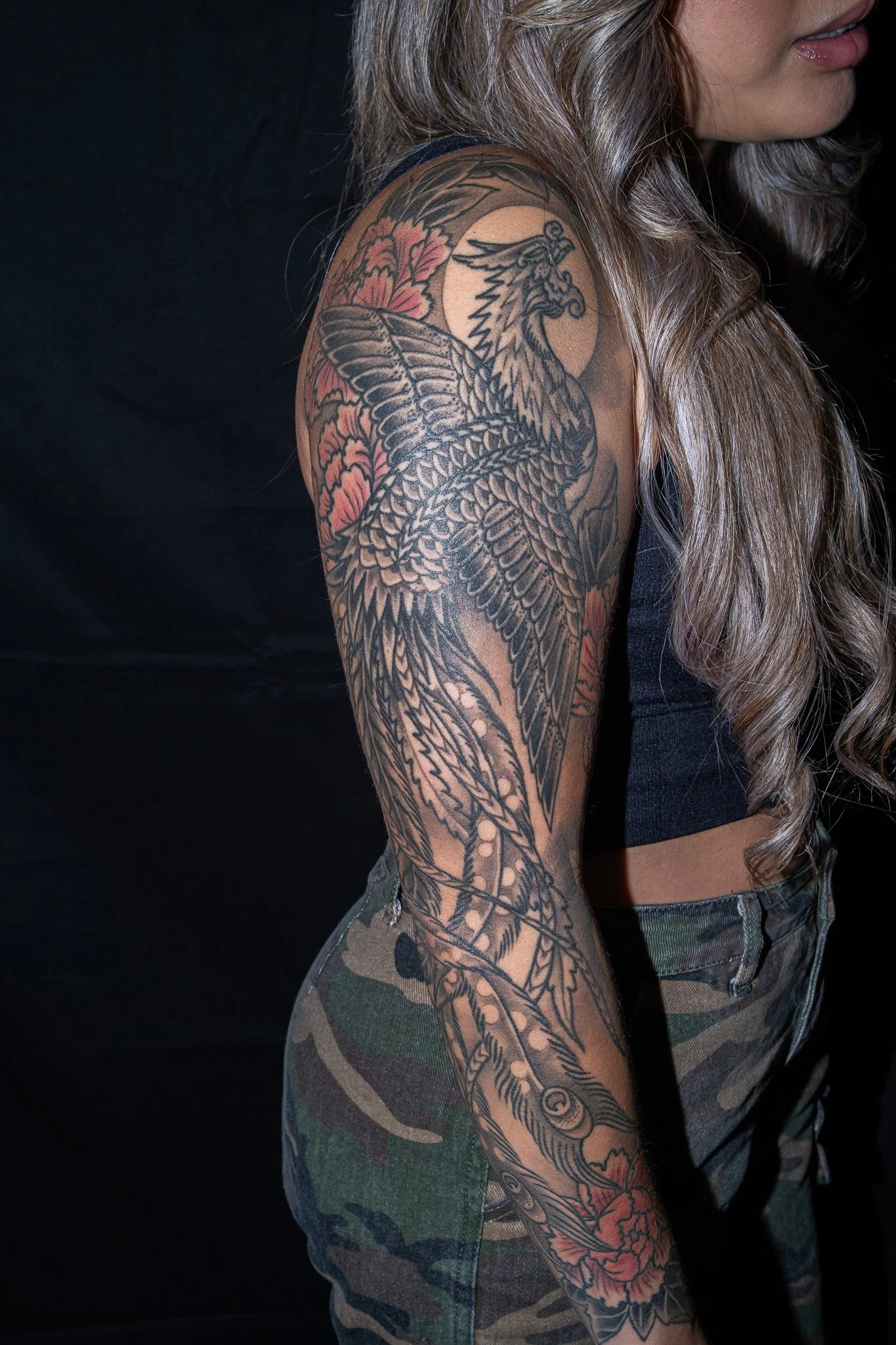 A woman with long, wavy hair and lipstick is showing her large arm tattoo of a phoenix with detailed feathers, flowers, and a sun or moon in the background, wearing camouflage shorts. Tattoo by Joe Haasch. 