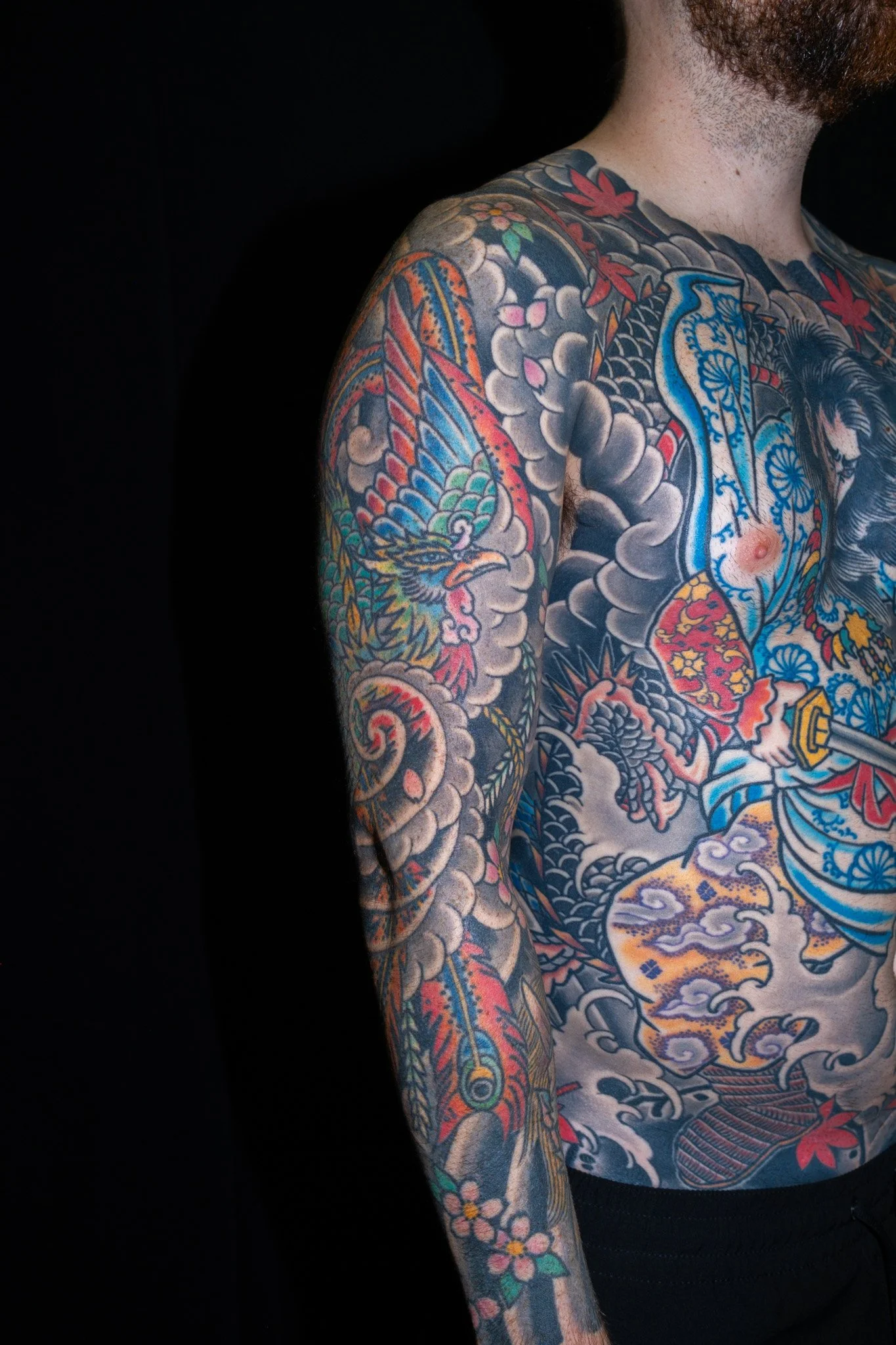 Partial view of a person with extensive colorful tattoos on their shoulder, chest, and arm. The tattoo features a Japanese design with a dragon, clouds, flowers, and traditional motifs.