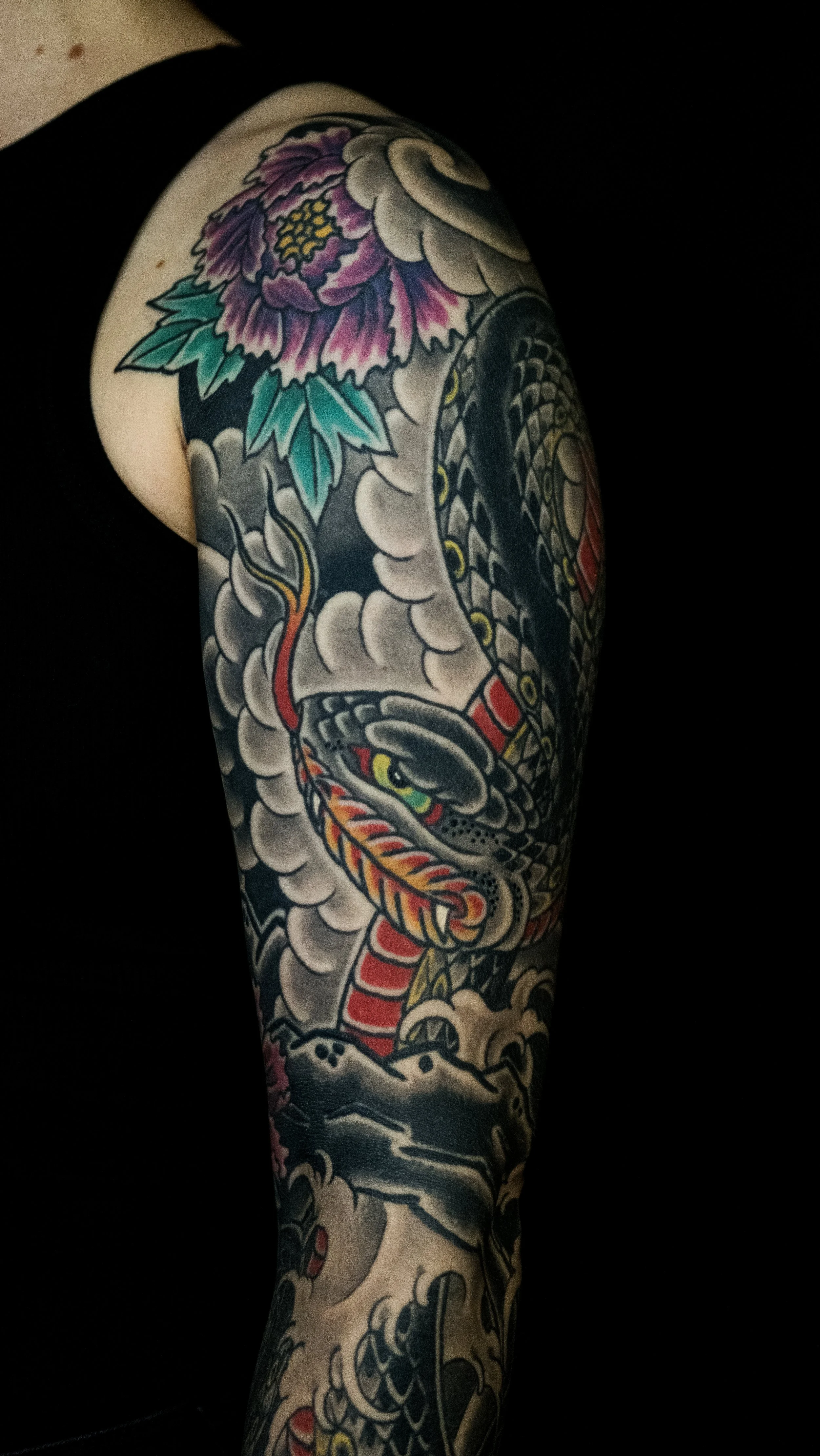 Tattoo sleeve on arm featuring a traditional Japanese dragon with detailed scales, flowing clouds, and a large peony flower with purple and pink petals and green leaves. Tattoo by Joe Haasch. 